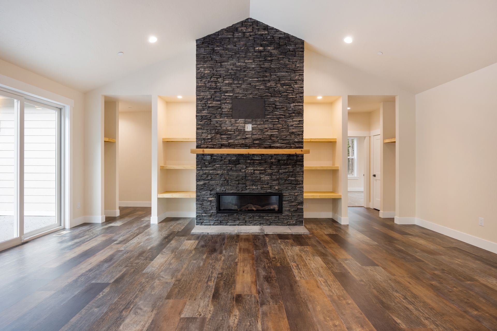 Empty living room with dark stone fireplace, wood floors, and built-in shelves around a central wall