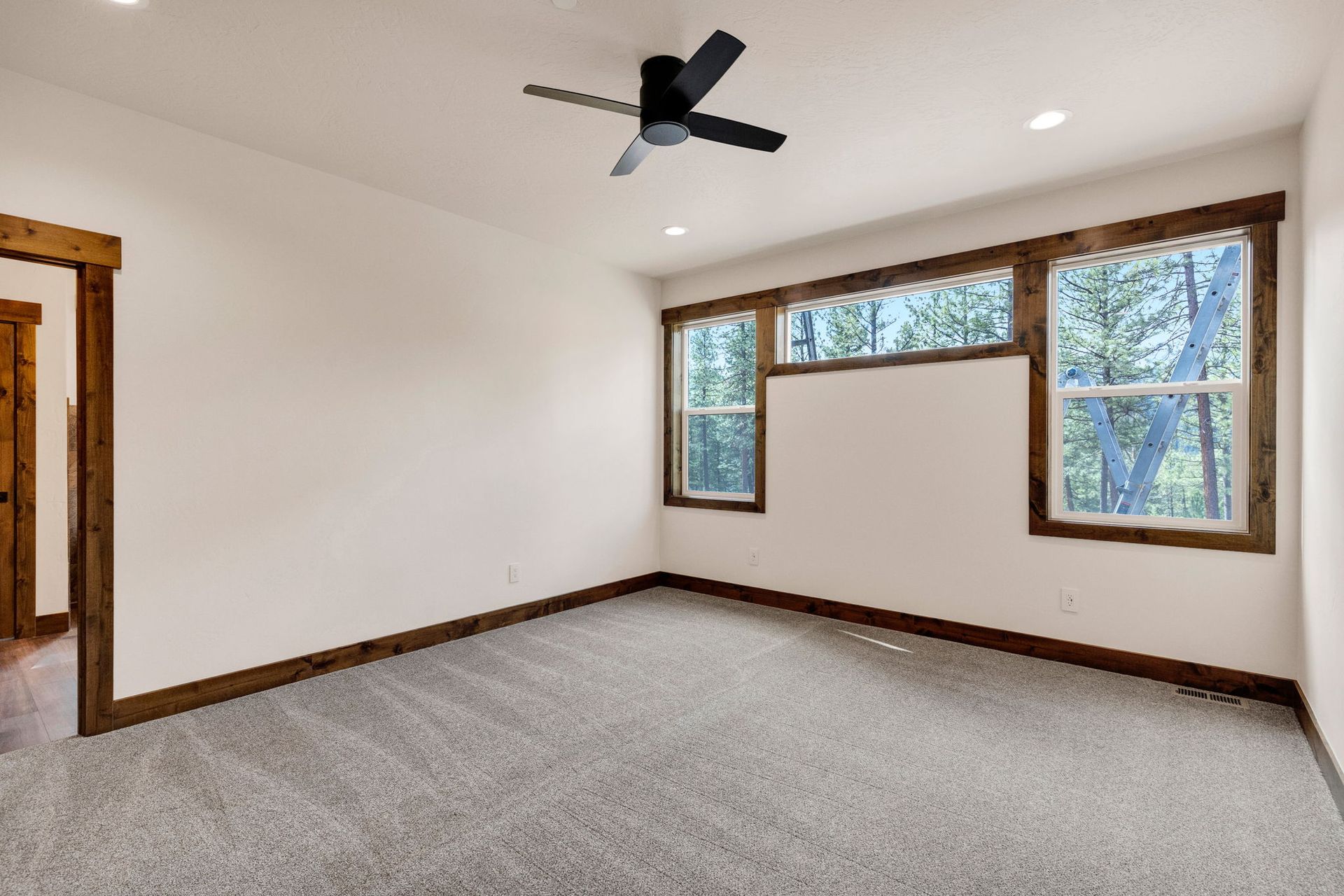 Empty carpeted bedroom with white walls, wood trim, ceiling fan, and large windows.