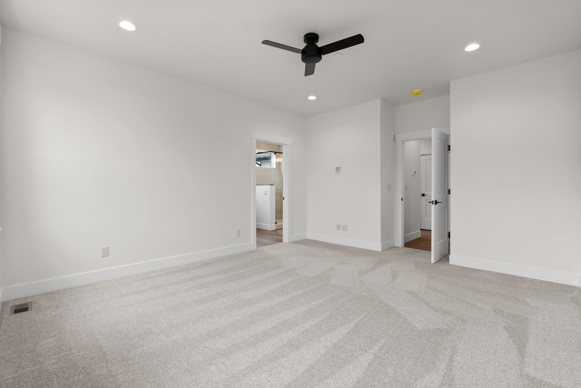 Empty white bedroom with carpet, ceiling fan, and open doorways to a bathroom and hallway