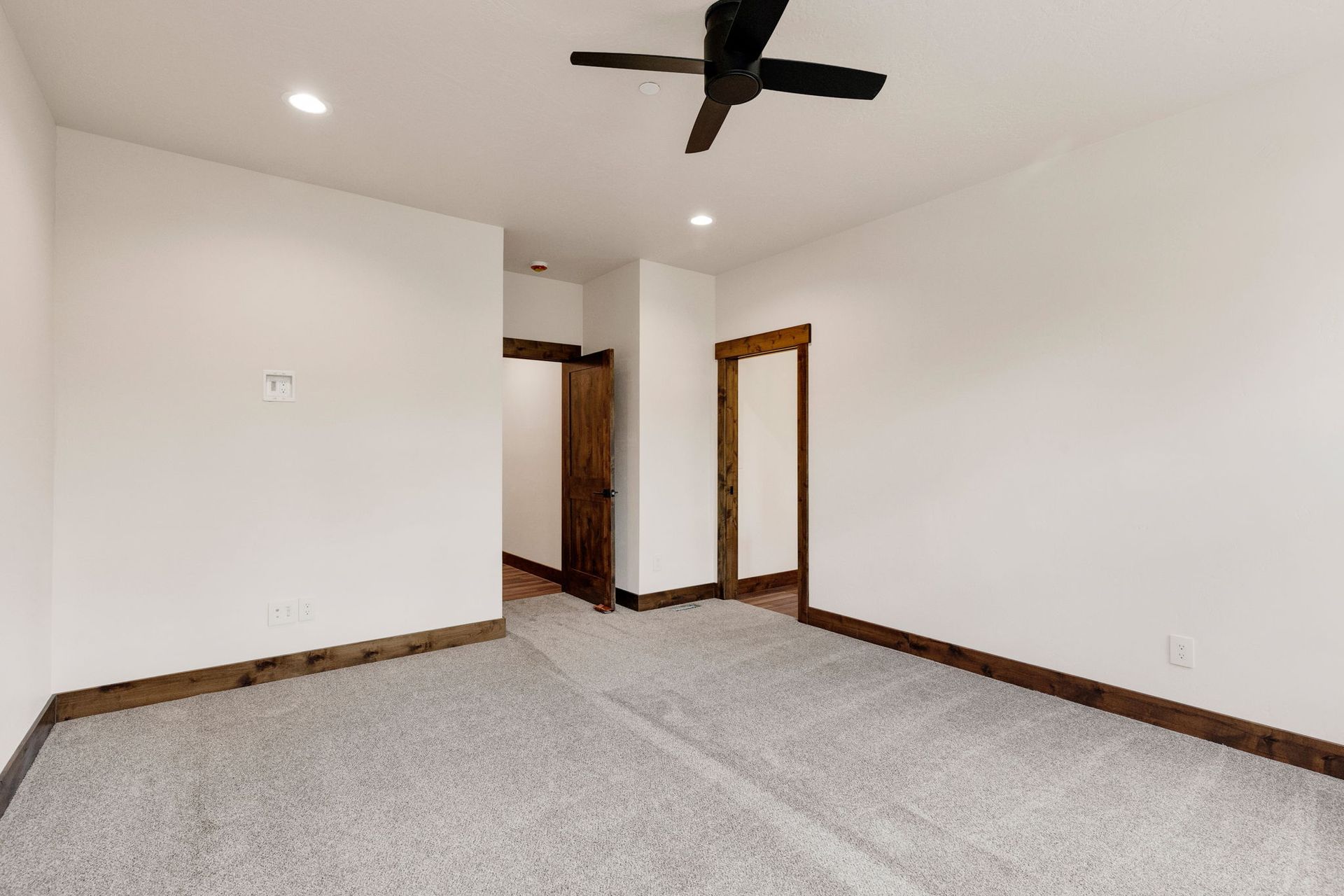 Empty carpeted bedroom with white walls, wooden trim, two doorways, and a black ceiling fan.