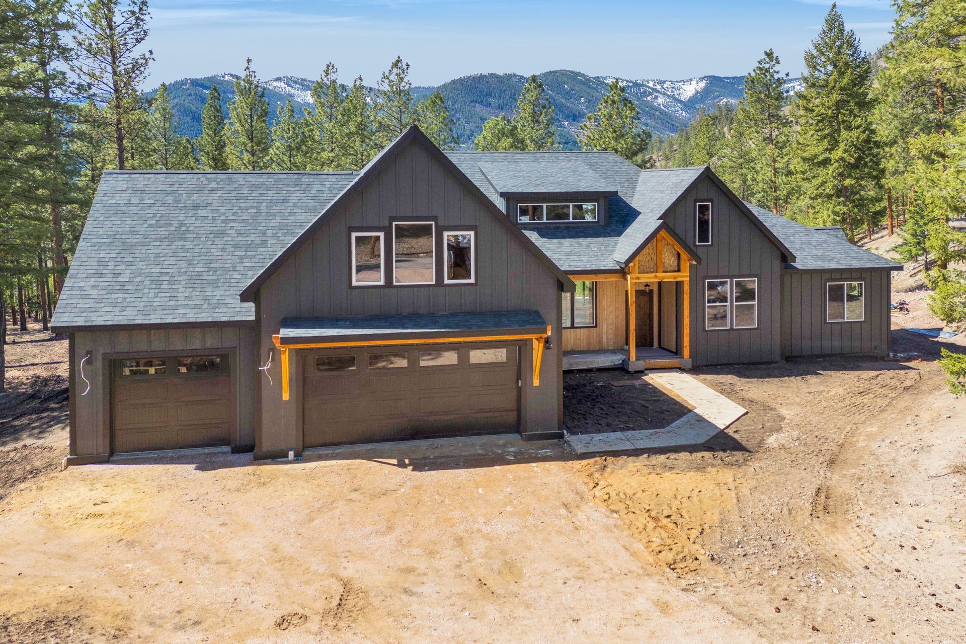 Modern dark-gray house with garage on a sunny wooded hillside, mountains in the background