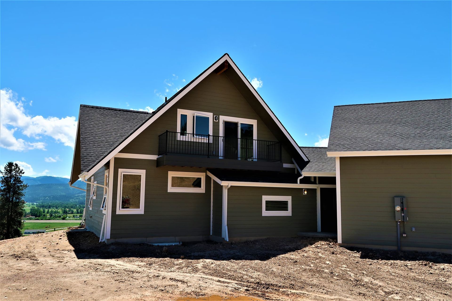 New house exterior with tan siding, dark roof, and dirt foreground under a blue sky