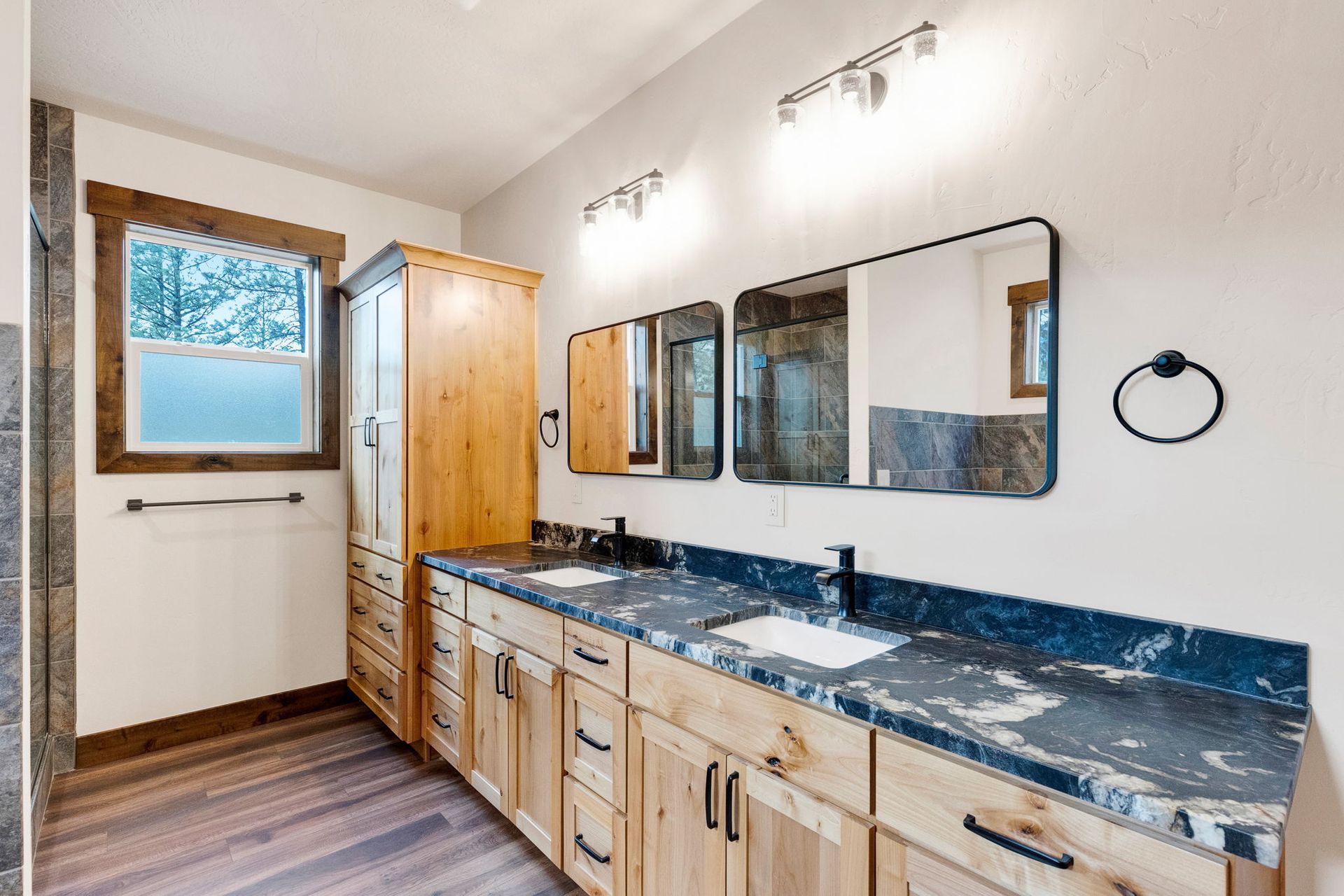Bathroom vanity with double mirrors, wood cabinets, black countertop, and window beside the sink