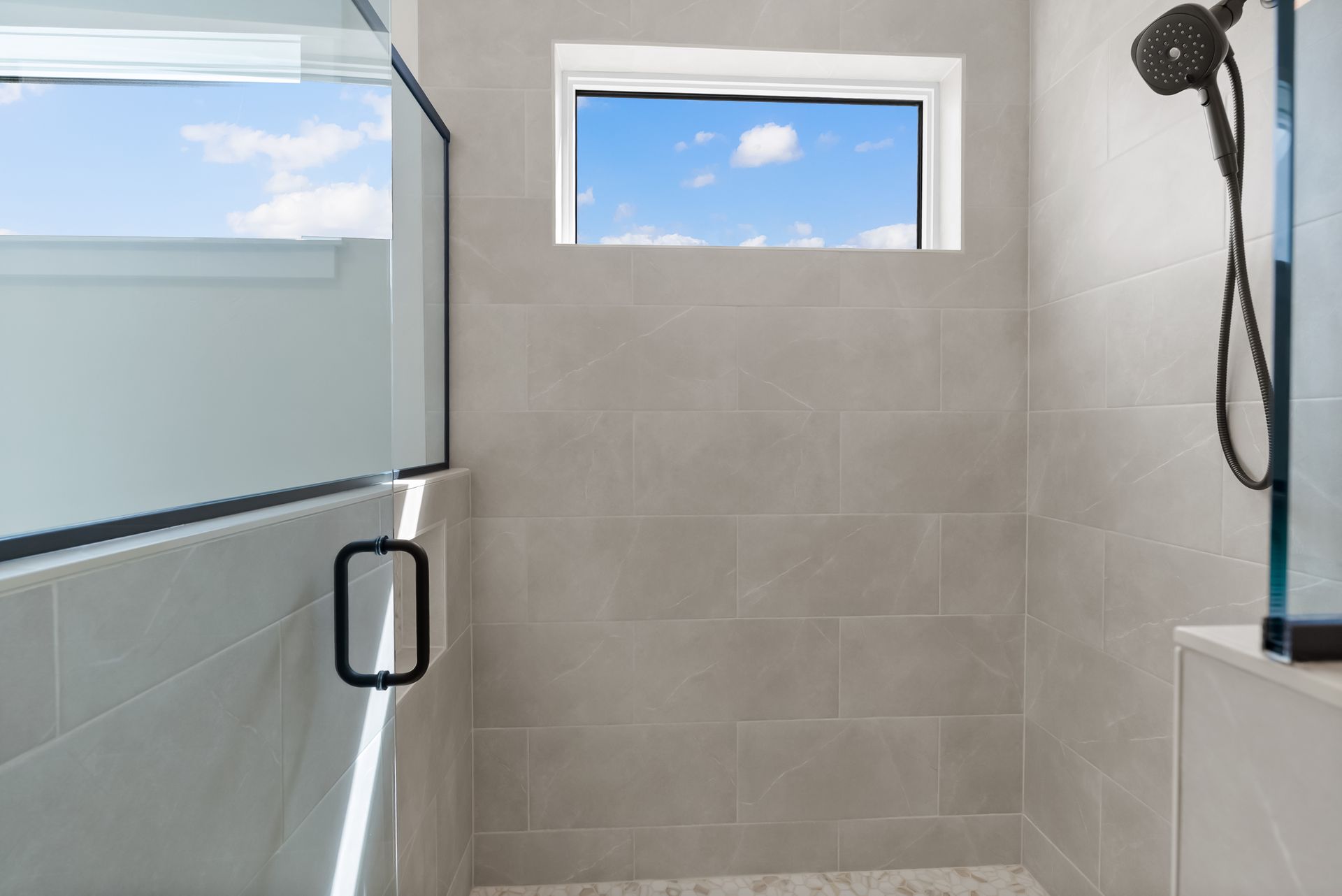 Modern bathroom shower with glass door, tiled walls, small window, and handheld showerhead.