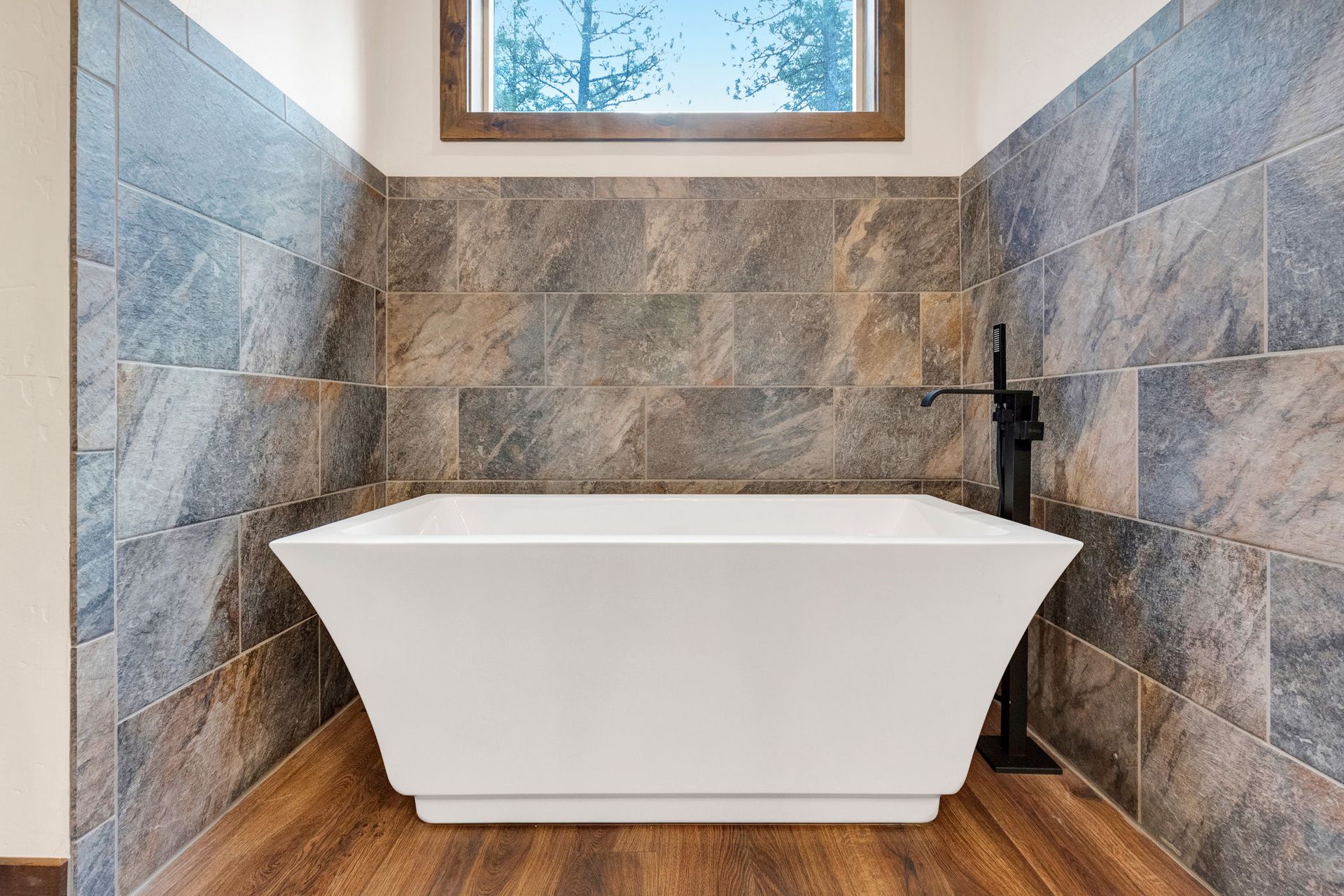 Modern bathroom with a white freestanding tub, stone tile walls, wood floor, and small window above.