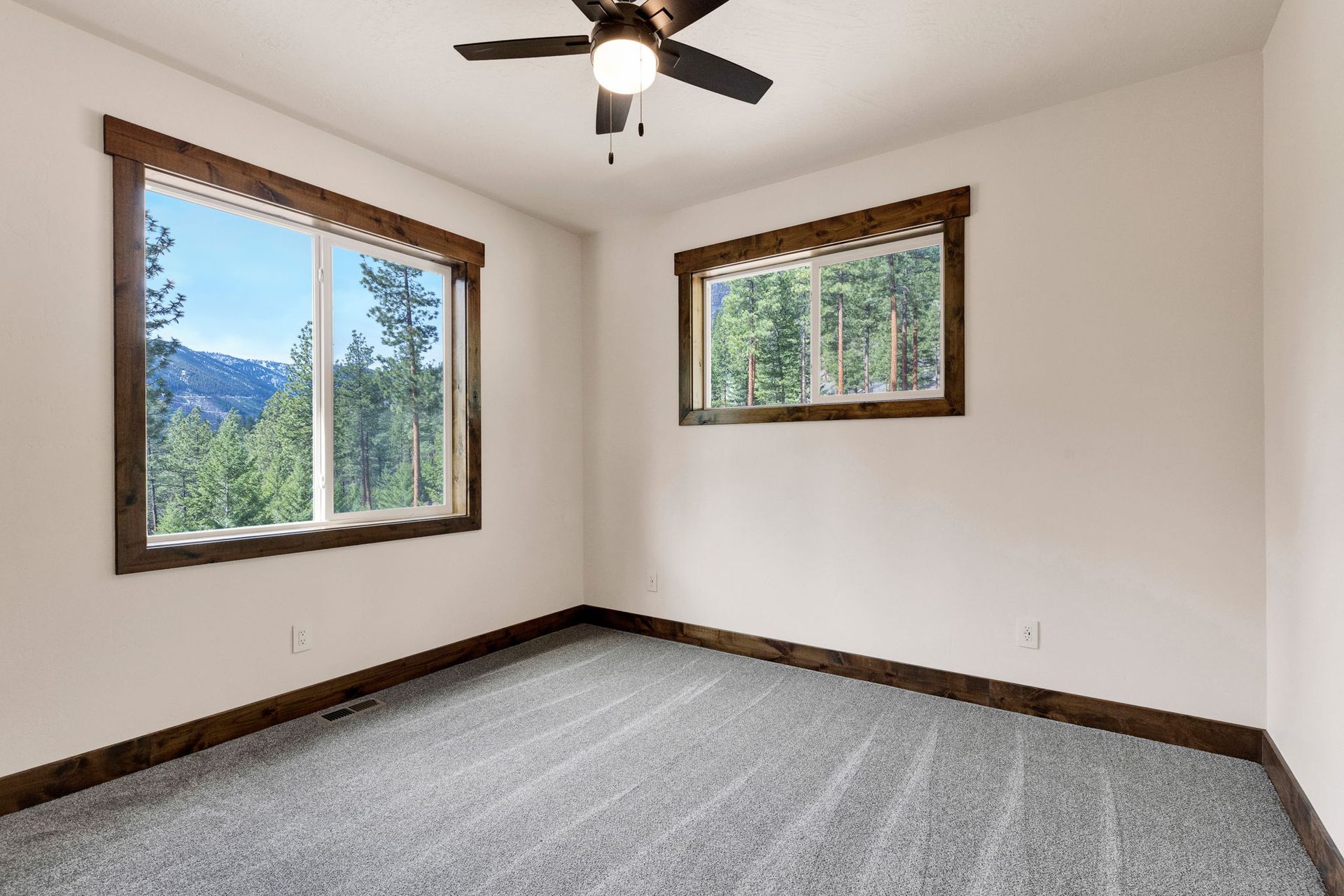 Empty carpeted room with beige walls, two windows, wood trim, and a ceiling fan.