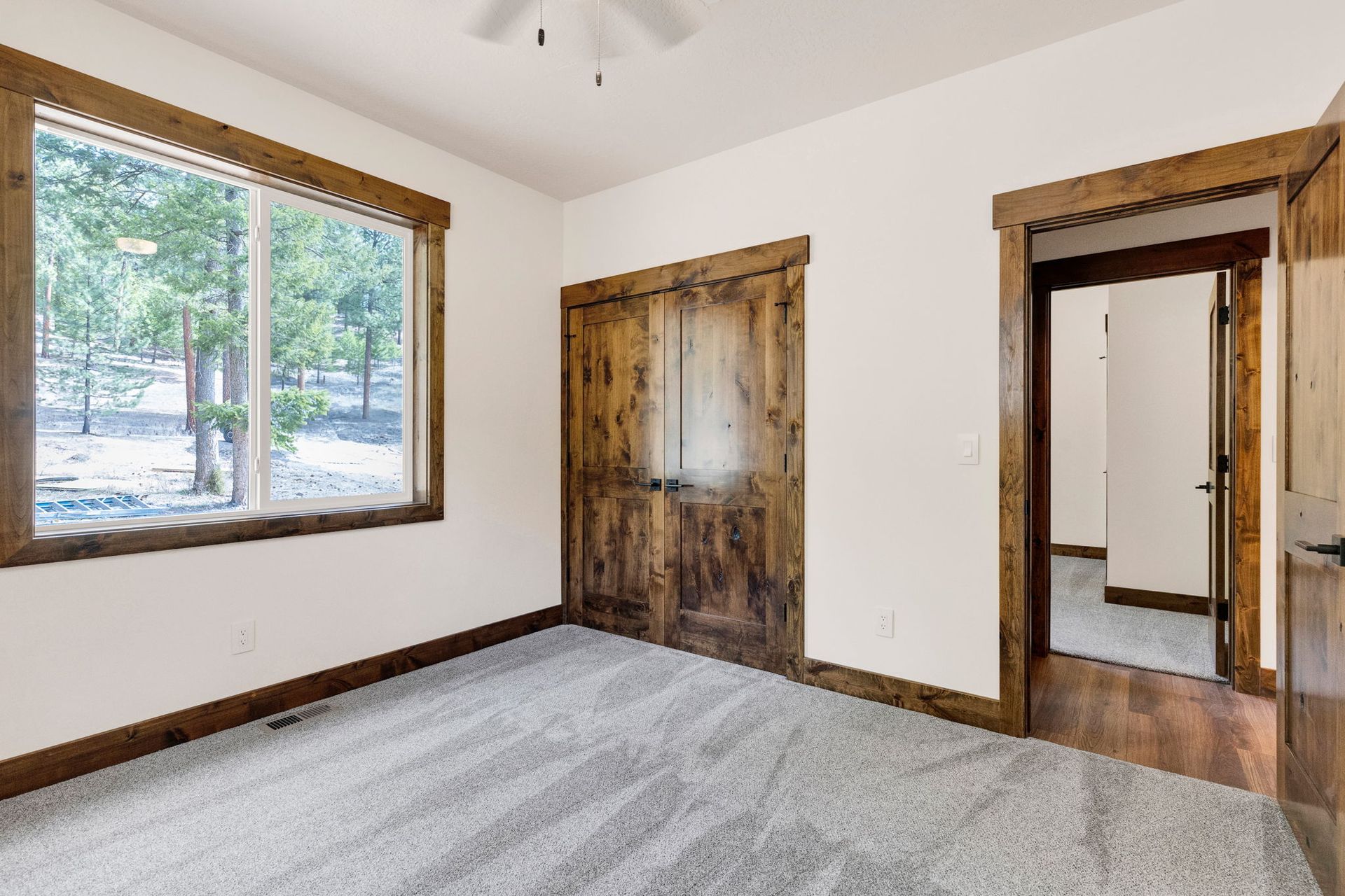 Empty carpeted room with rustic wood-trimmed windows and doors, white walls, and natural light.