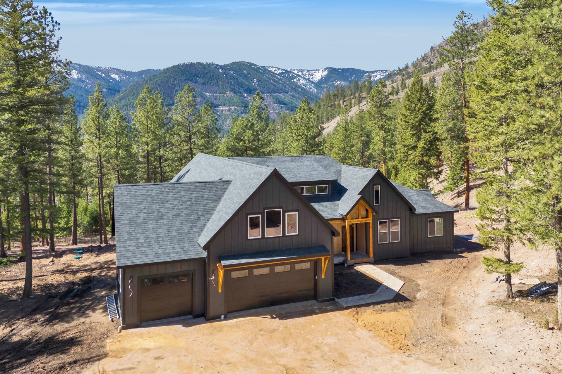 Modern dark-gray cabin with garage in a pine forest, backed by mountains under a clear sky