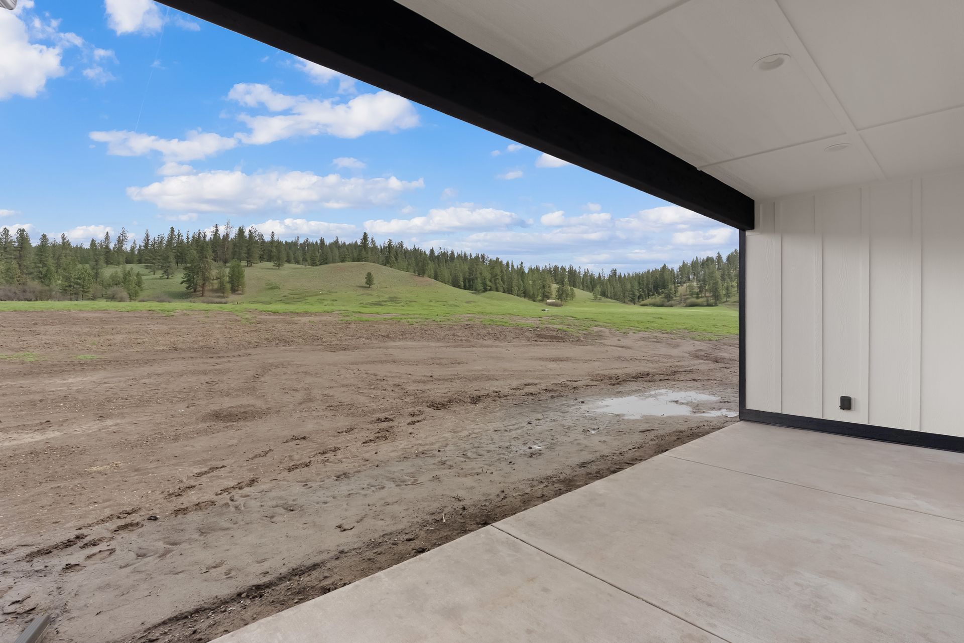 Covered patio overlooking a bare dirt lot and green trees under a cloudy blue sky