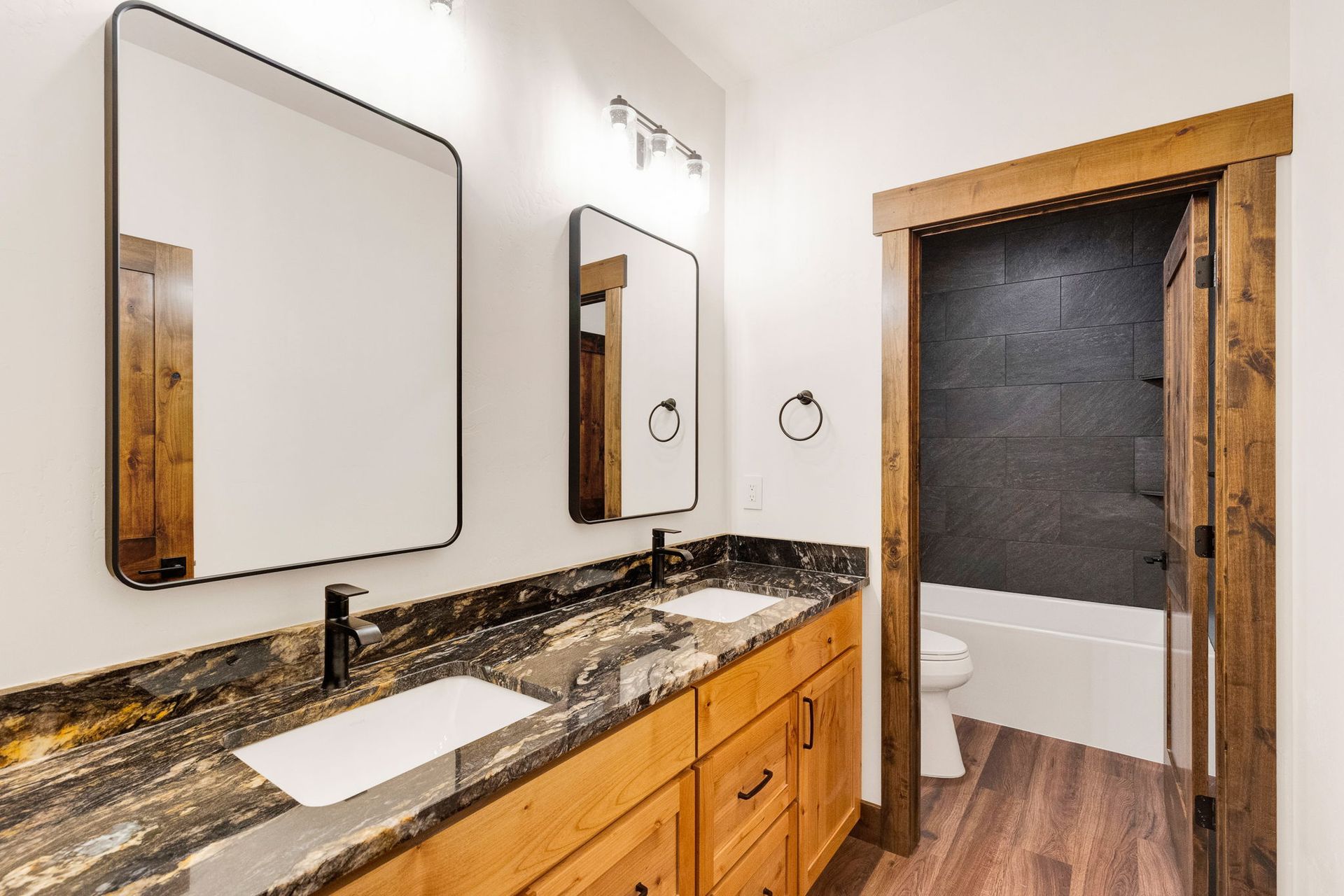 Modern bathroom vanity with black granite countertop, wood cabinets, and mirrors beside a shower doorway