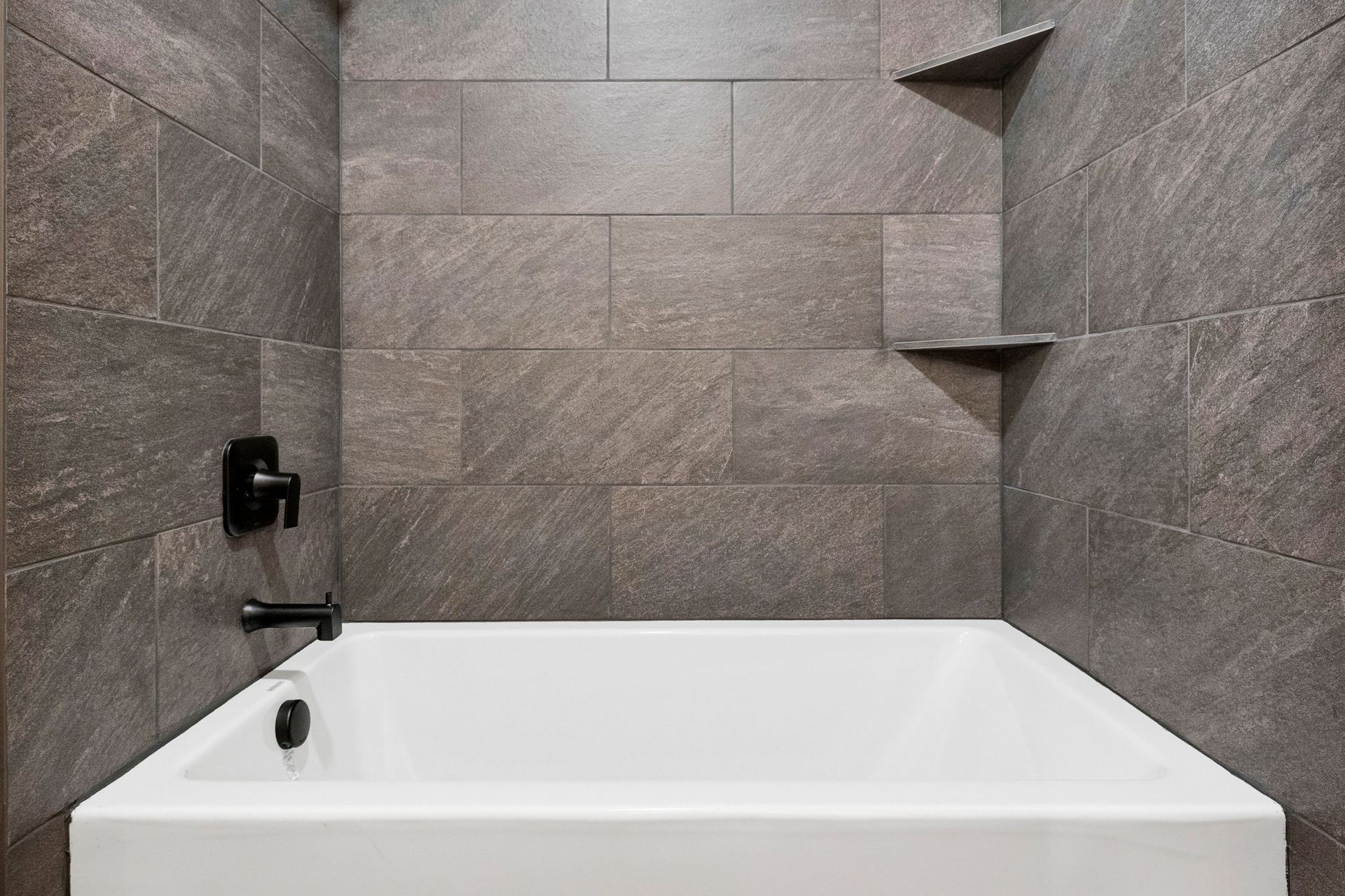 Modern bathroom with a white bathtub, gray tiled walls, black fixtures, and built-in corner shelves