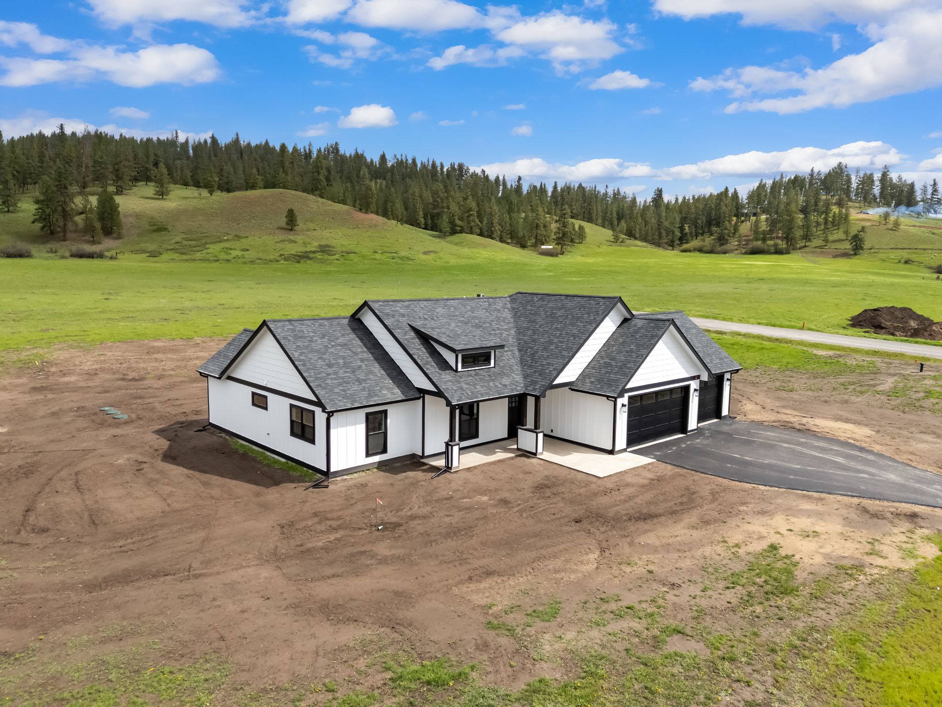 New white house with dark roof on a dirt lot beside green fields under a blue sky