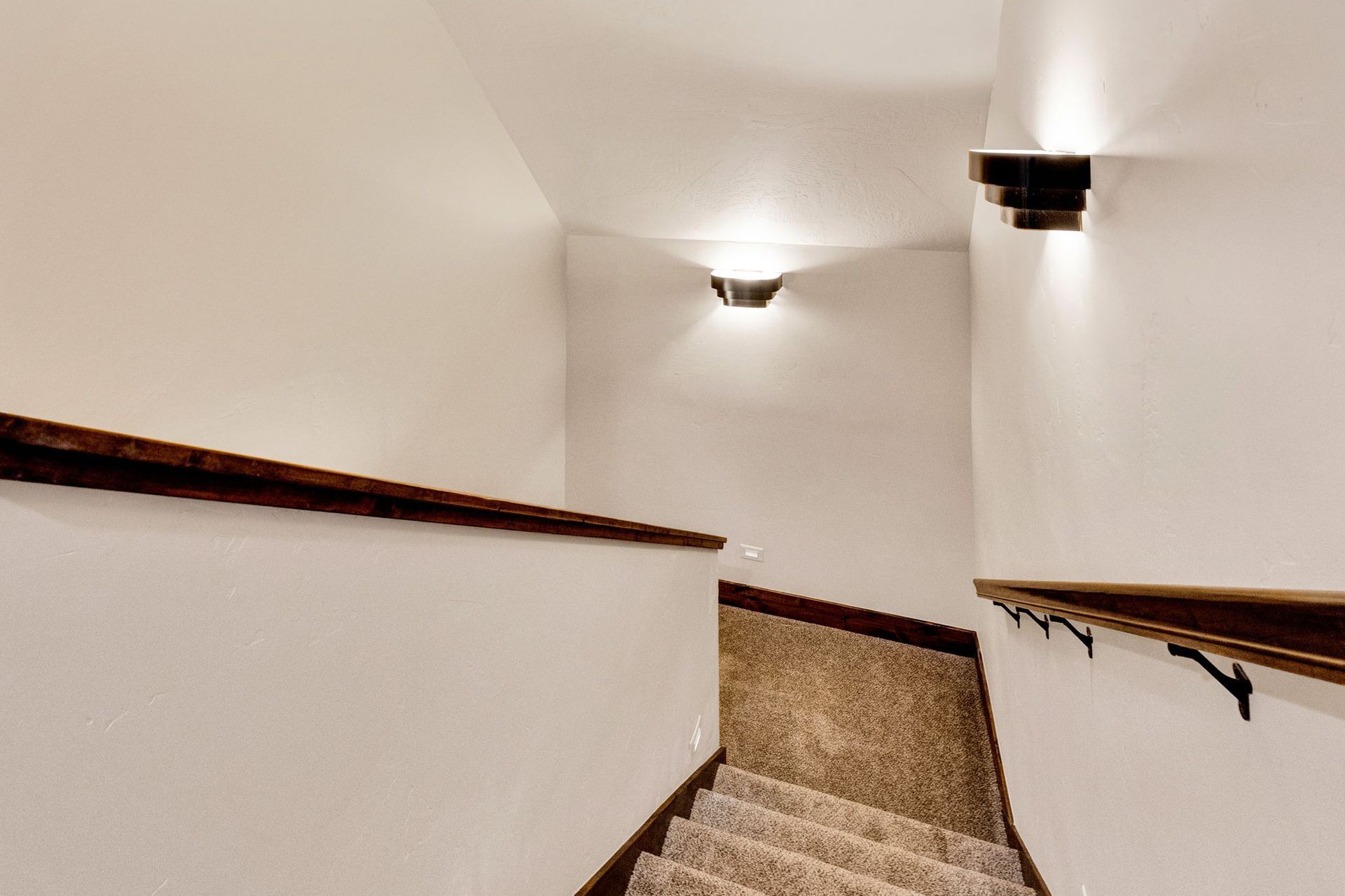 Narrow stairwell with beige walls, wooden handrails, and carpeted steps descending to a lower floor