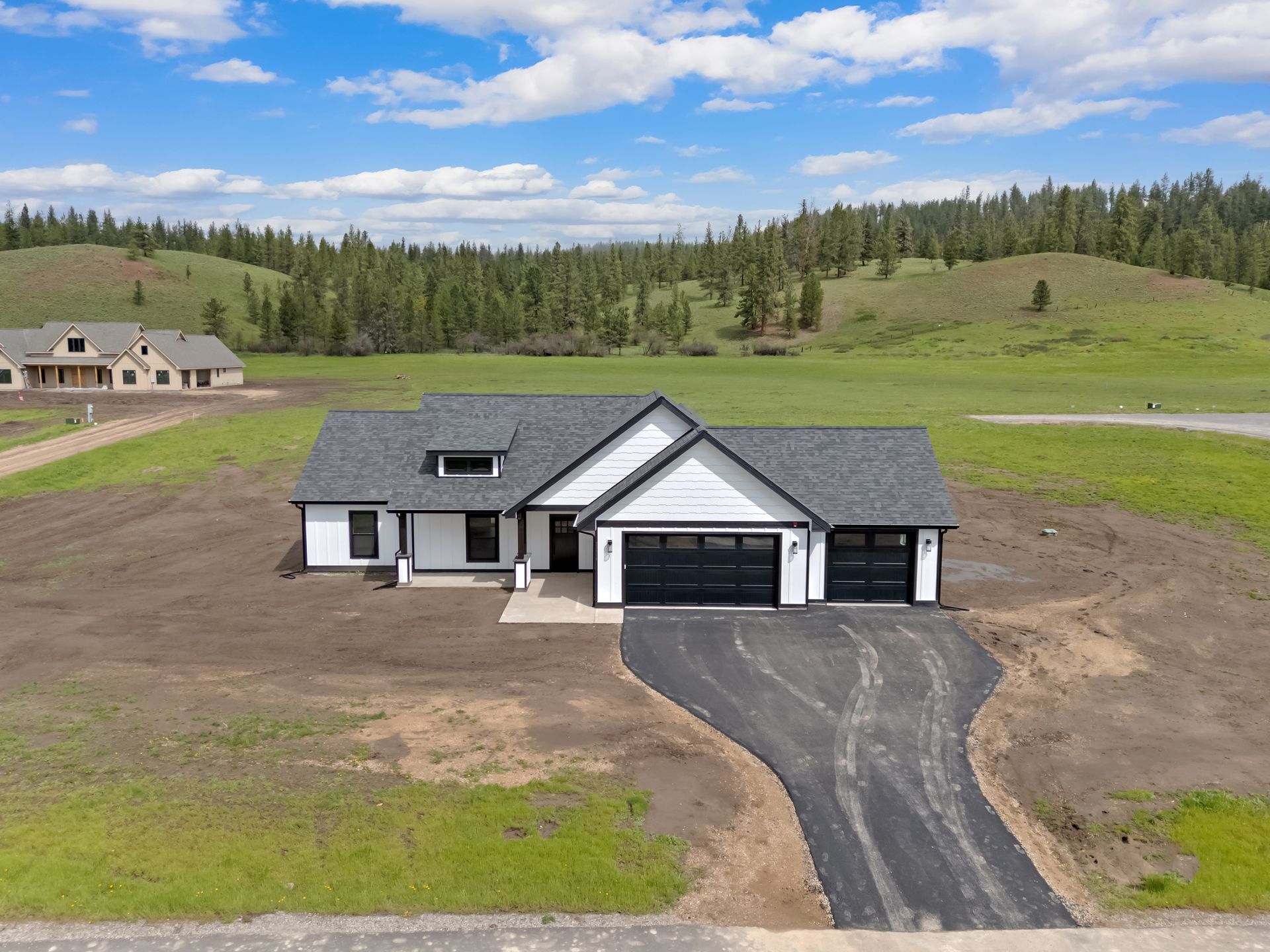 Single-story gray house with black roof and long driveway in a rural field under a blue sky