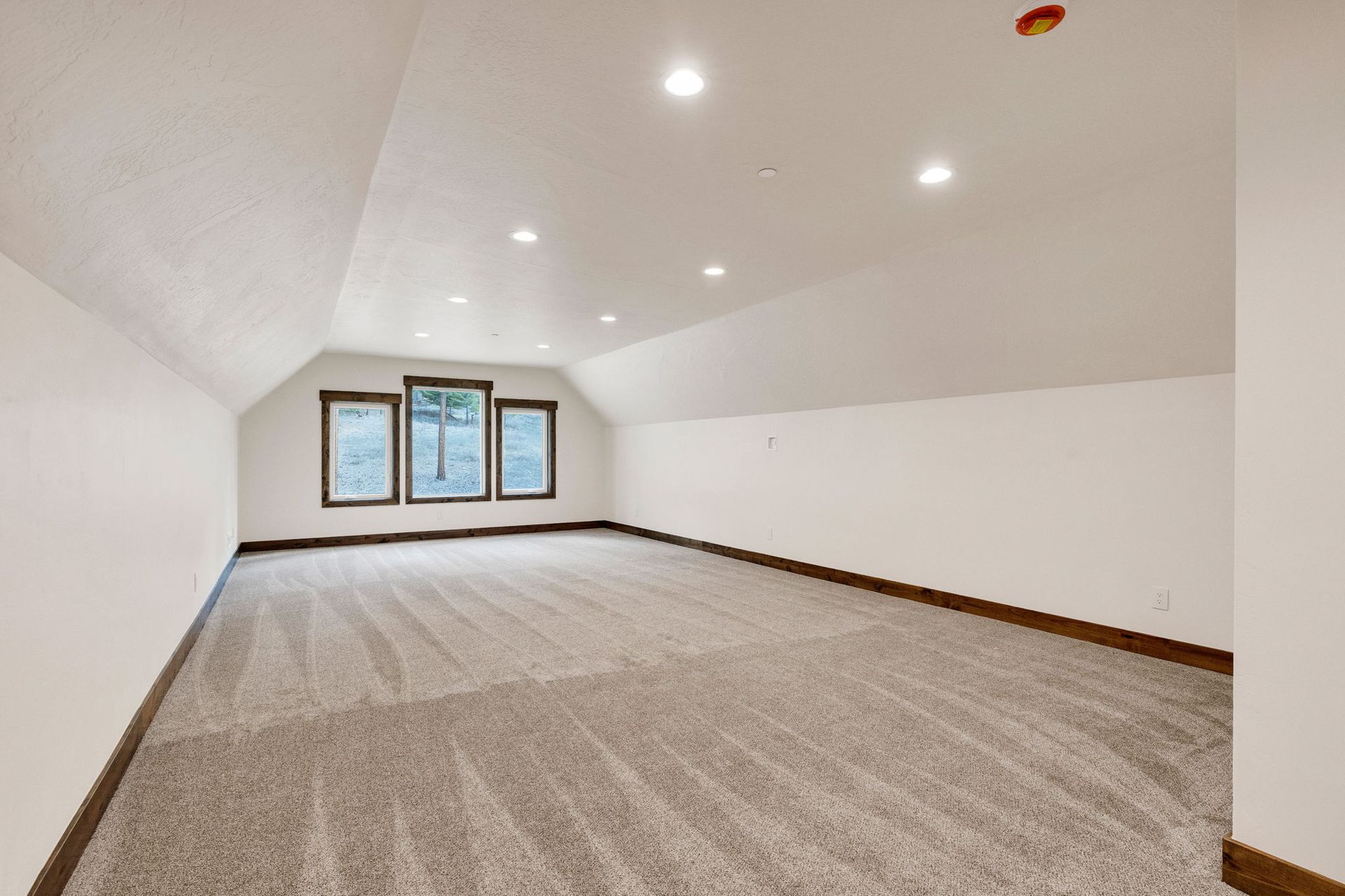 Empty carpeted attic room with white sloped ceilings, recessed lights, and three windows.