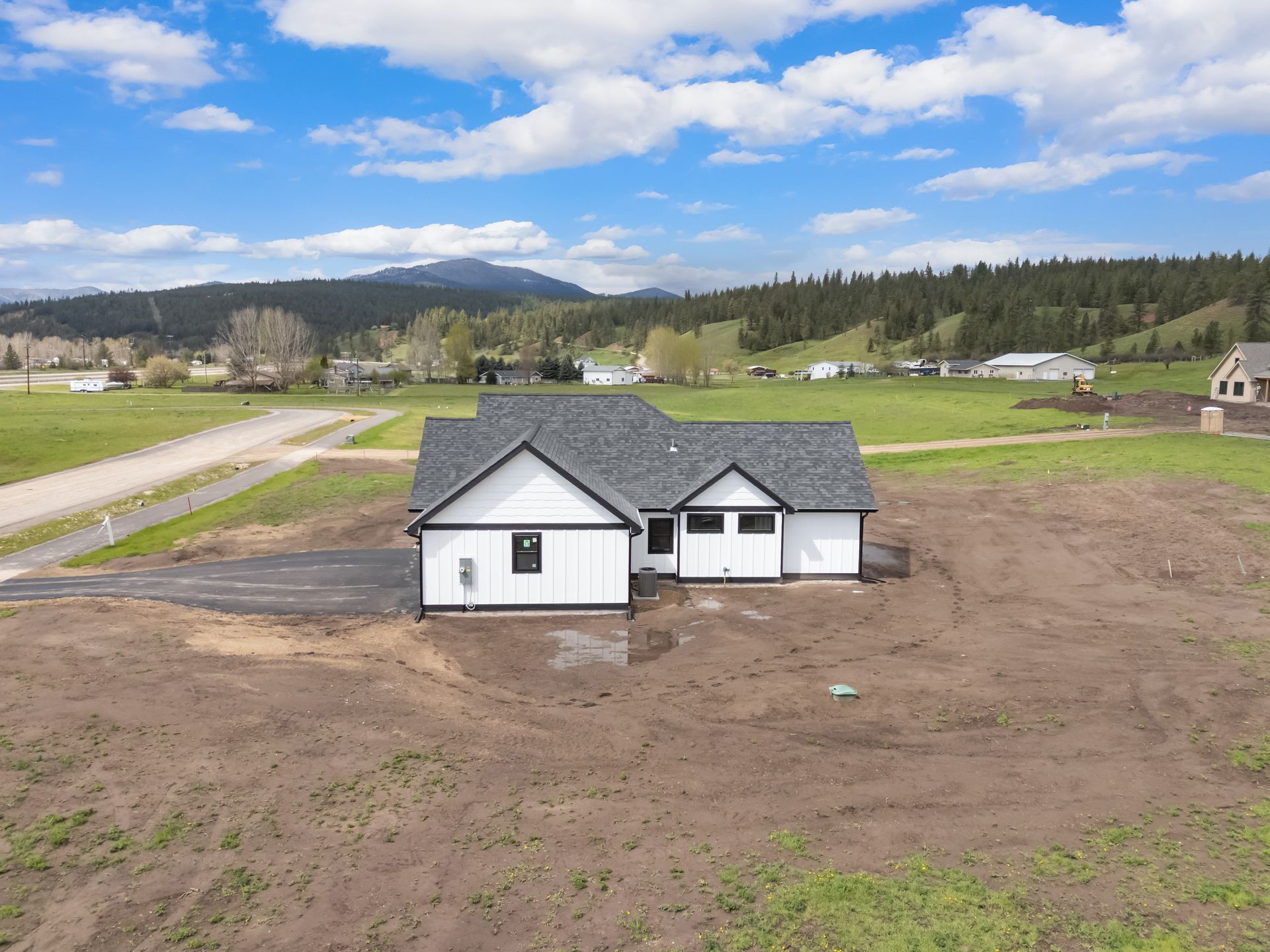 Small white house with gray roof on a dirt lot, with fields, road, and mountains under a blue sky.