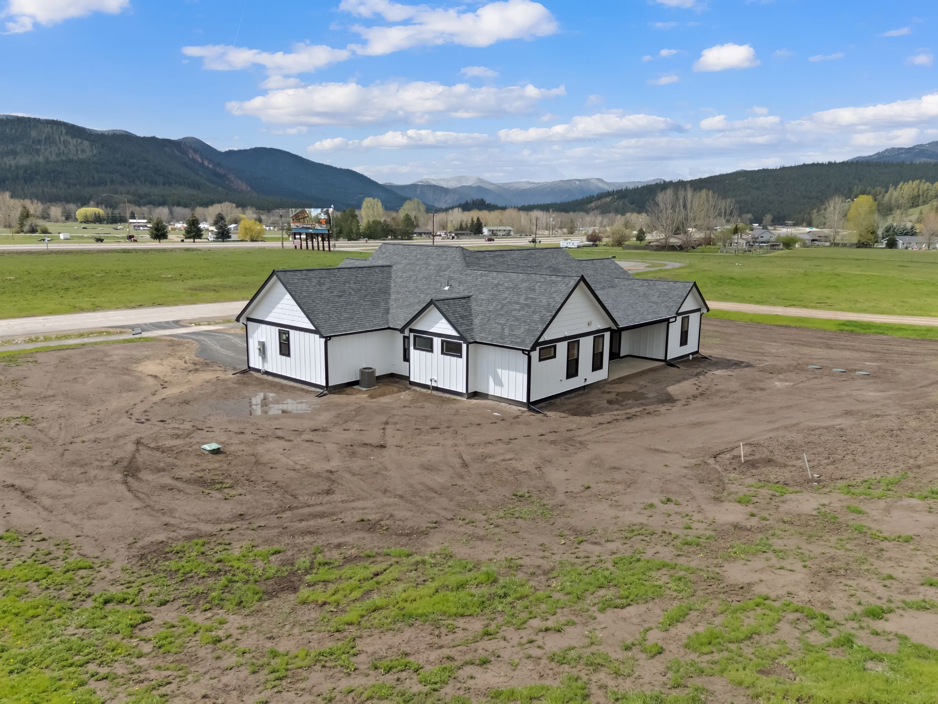 Small white house under construction on a dirt lot, with mountains and fields in the background.