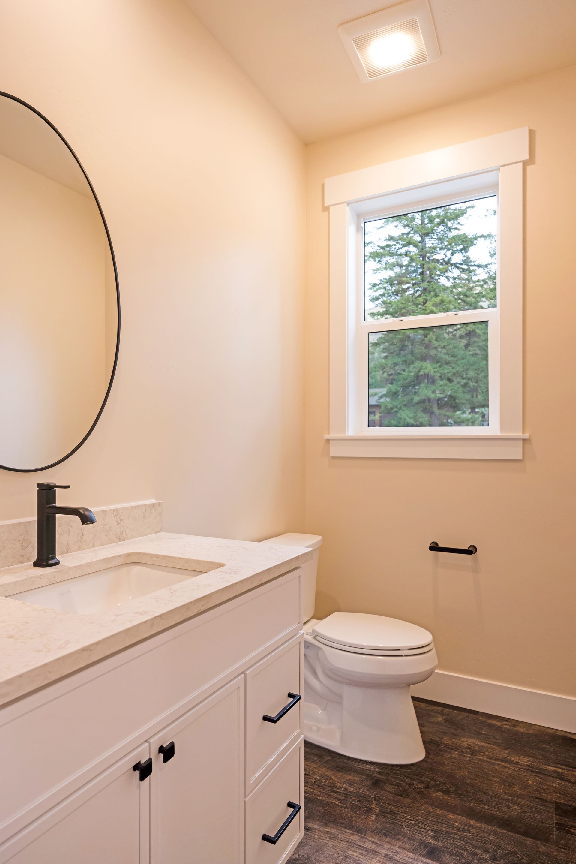 Small bathroom with white vanity, toilet, round mirror, and window above dark carpet.