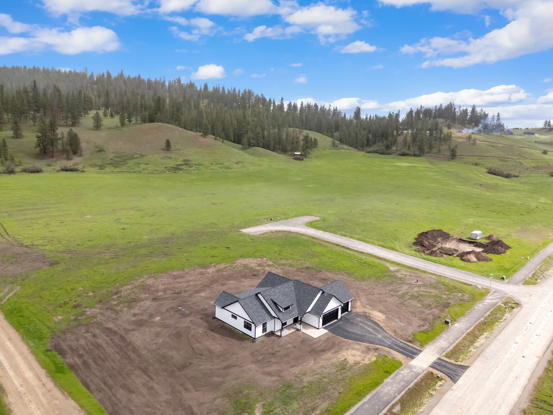 Aerial view of a house under construction on a grassy rural lot with dirt roads and forested hills beyond