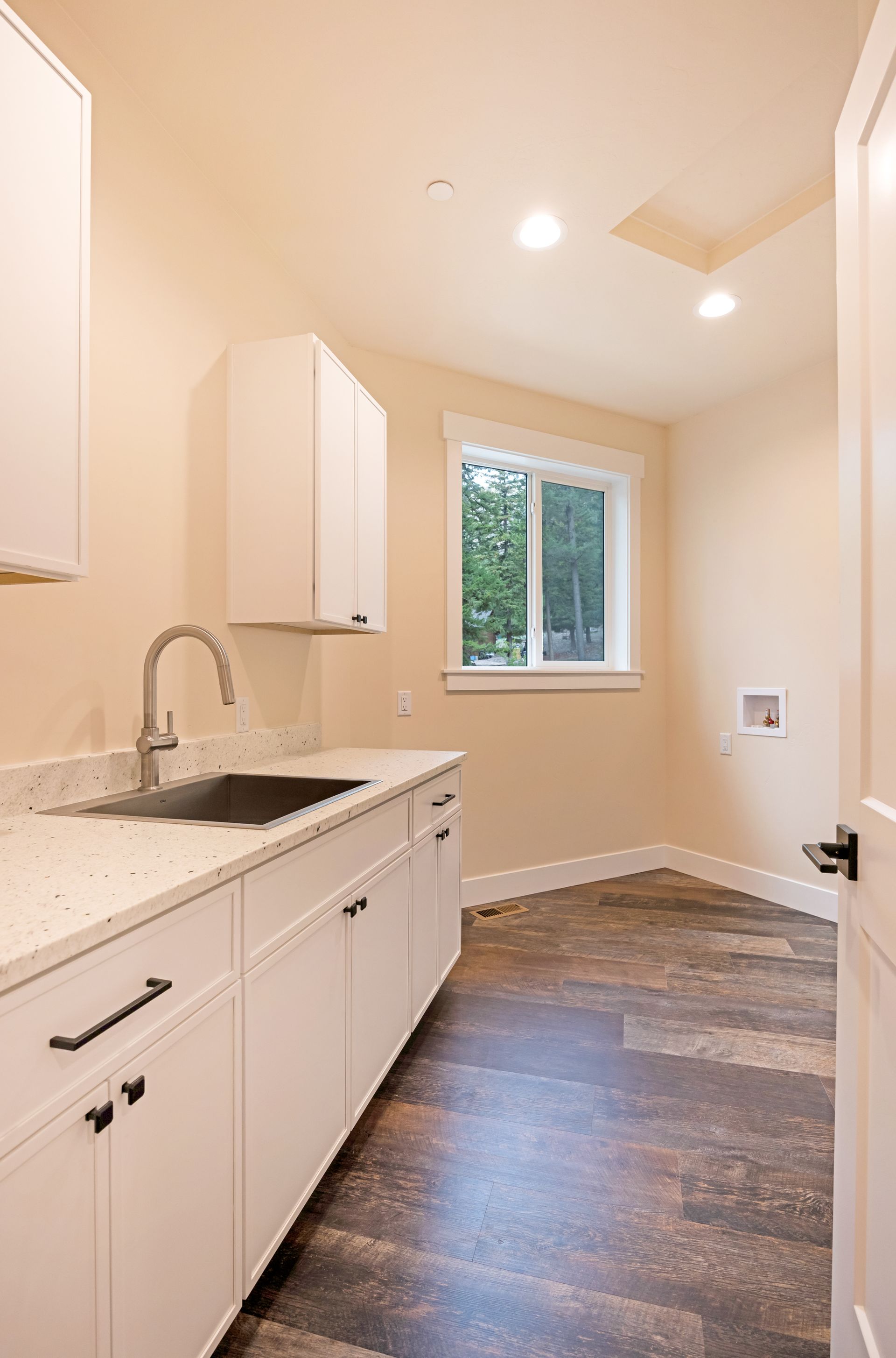 Bright laundry room with white cabinets, sink, window, and dark wood-look flooring