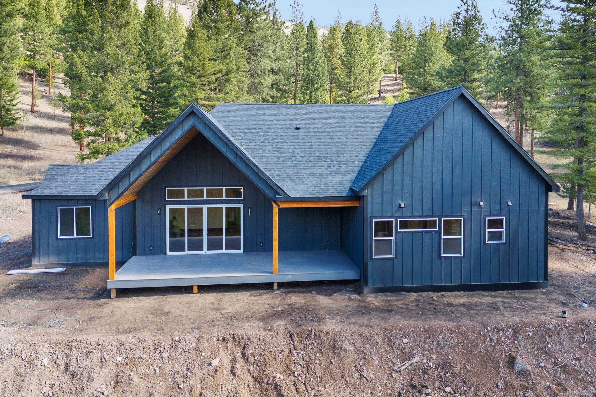Blue modern house under construction in a wooded area, with dark roof and unfinished dirt foreground