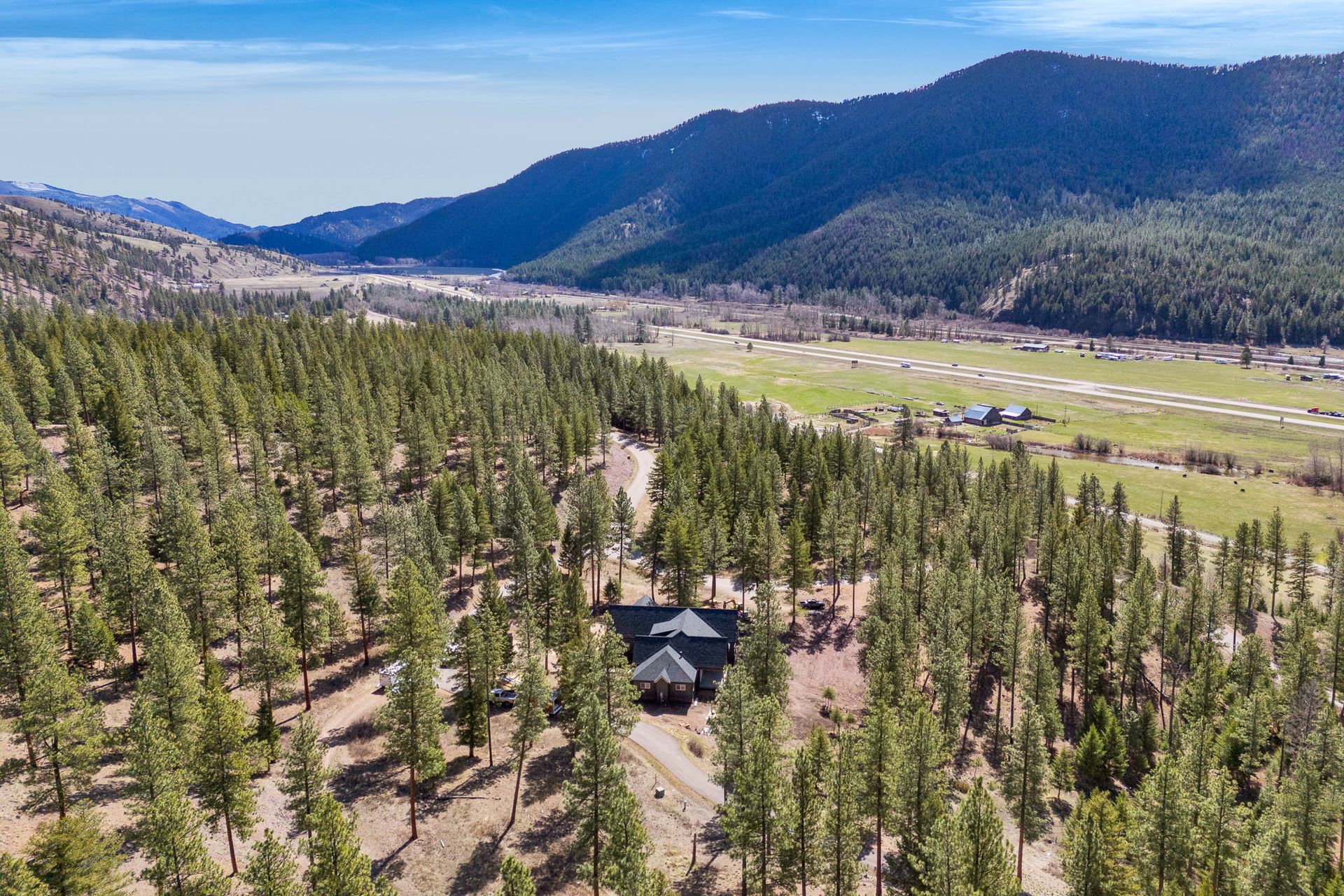 Aerial view of a forested mountain valley with a road, scattered cabins, and open meadow under a blue sky