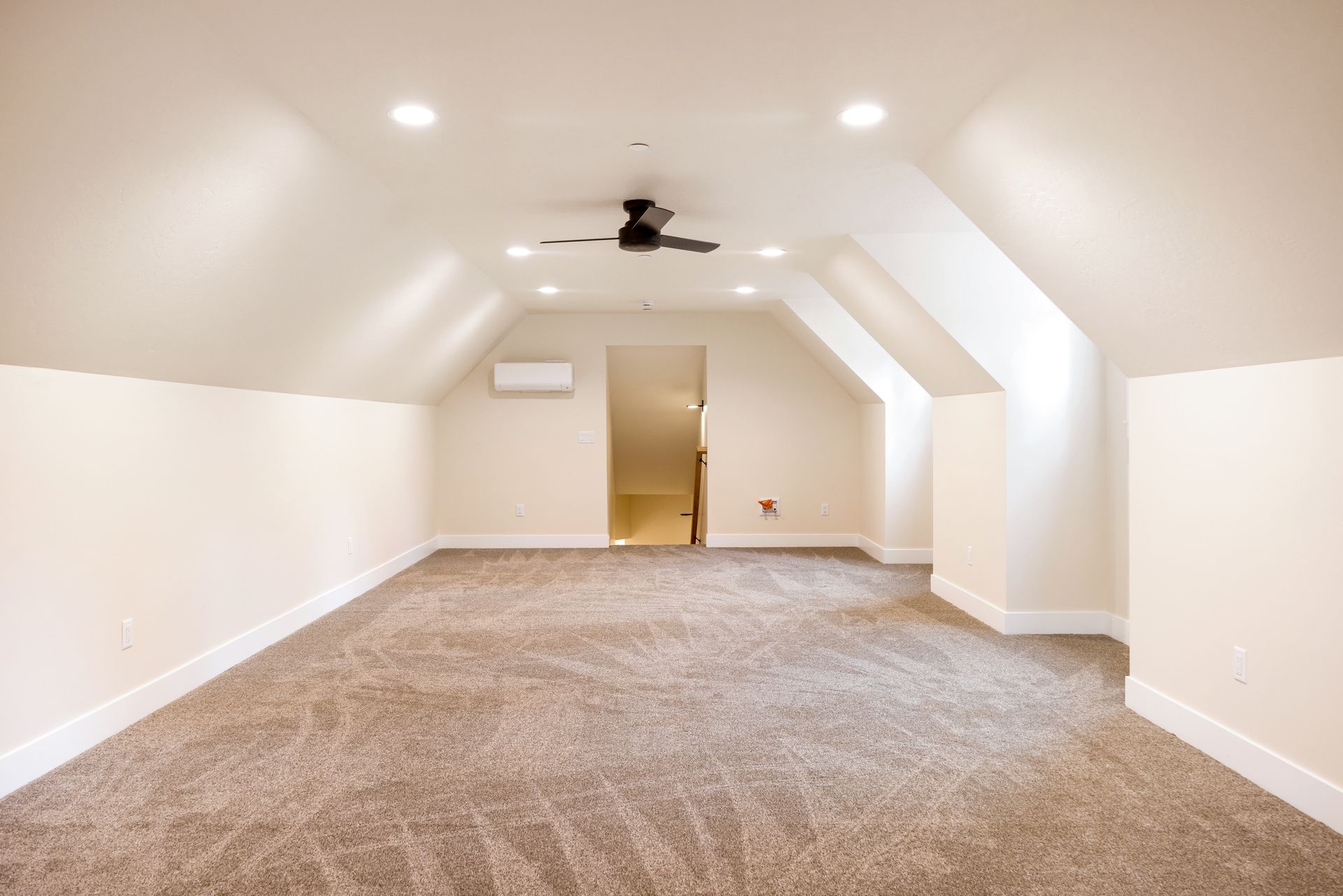 Empty carpeted attic room with white walls, sloped ceiling, and ceiling fan light.