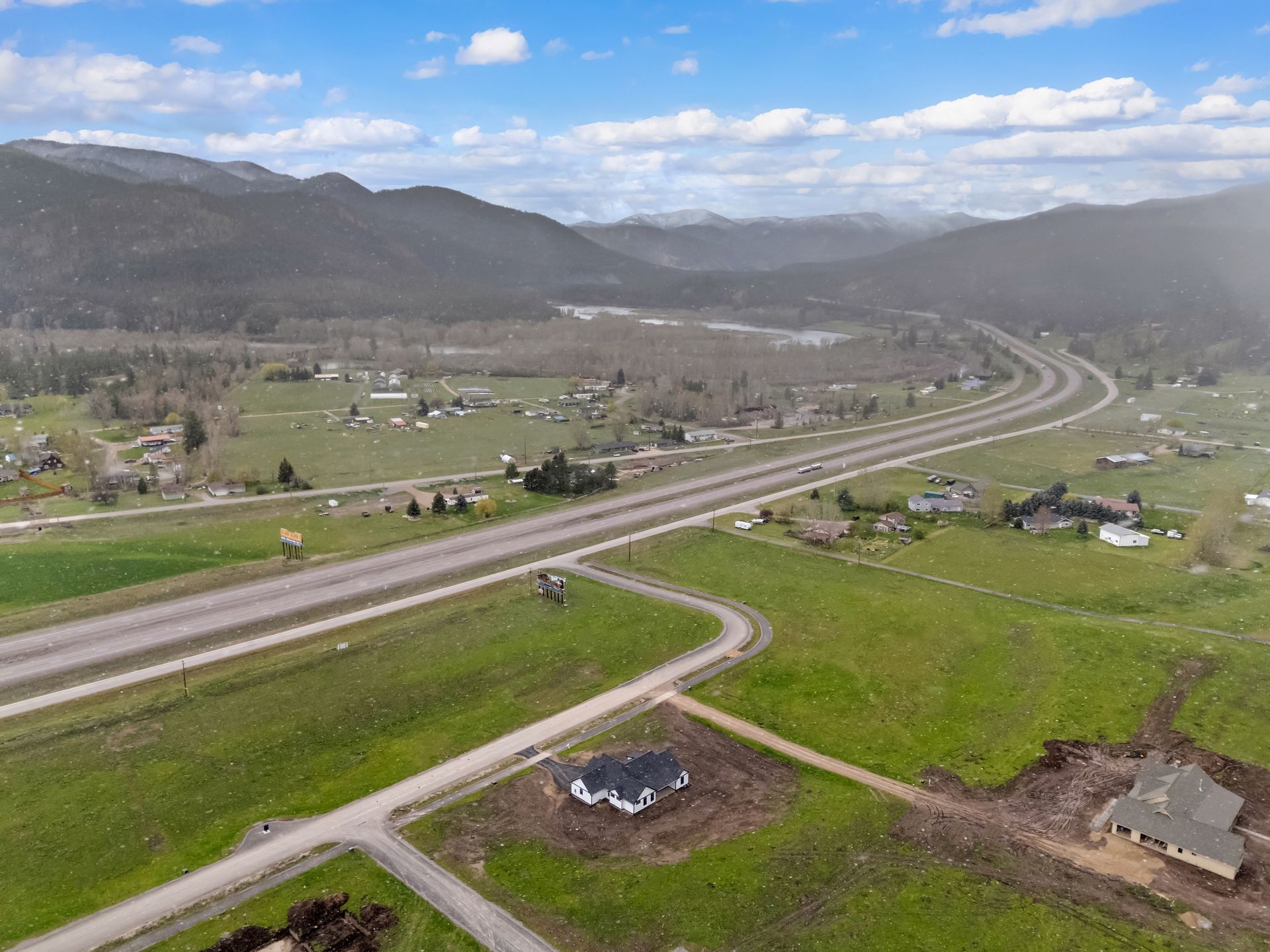 Aerial view of a highway cutting through a green valley with scattered houses and mountains under a blue sky