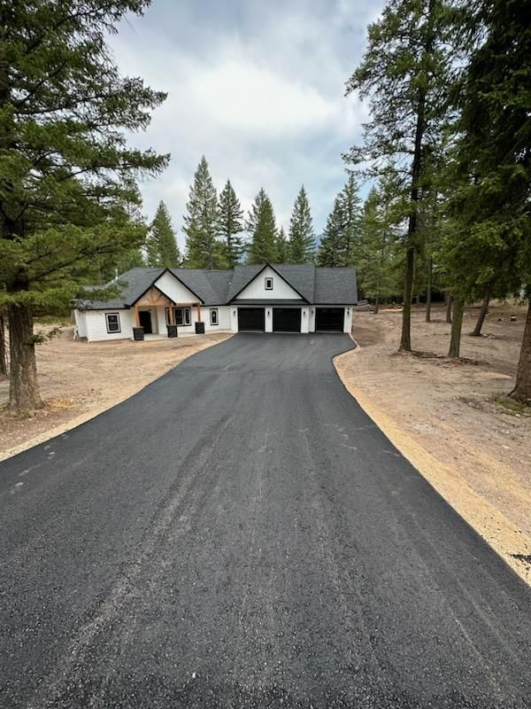 Long black driveway leading to a modern house with garages, surrounded by pine trees.