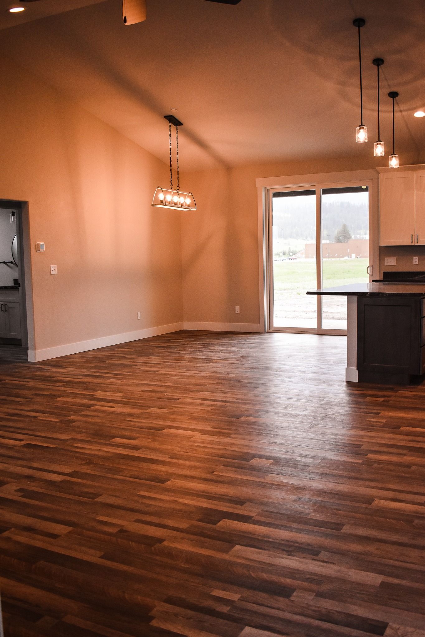 Empty modern living room with dark wood floors, beige walls, pendant lights, and sliding glass doors.
