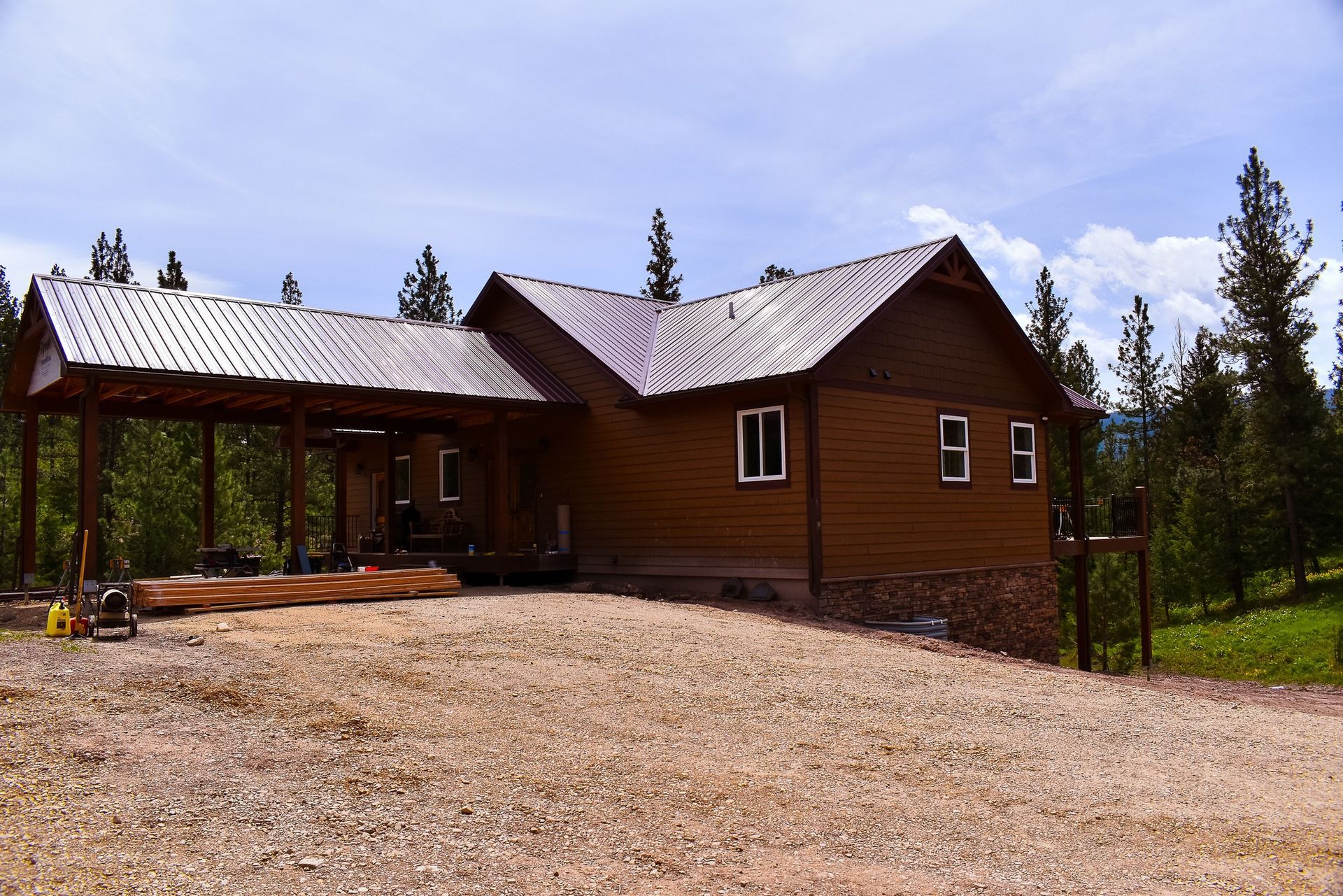 Wooden cabin with a metal roof on a gravel lot, surrounded by trees under a cloudy sky