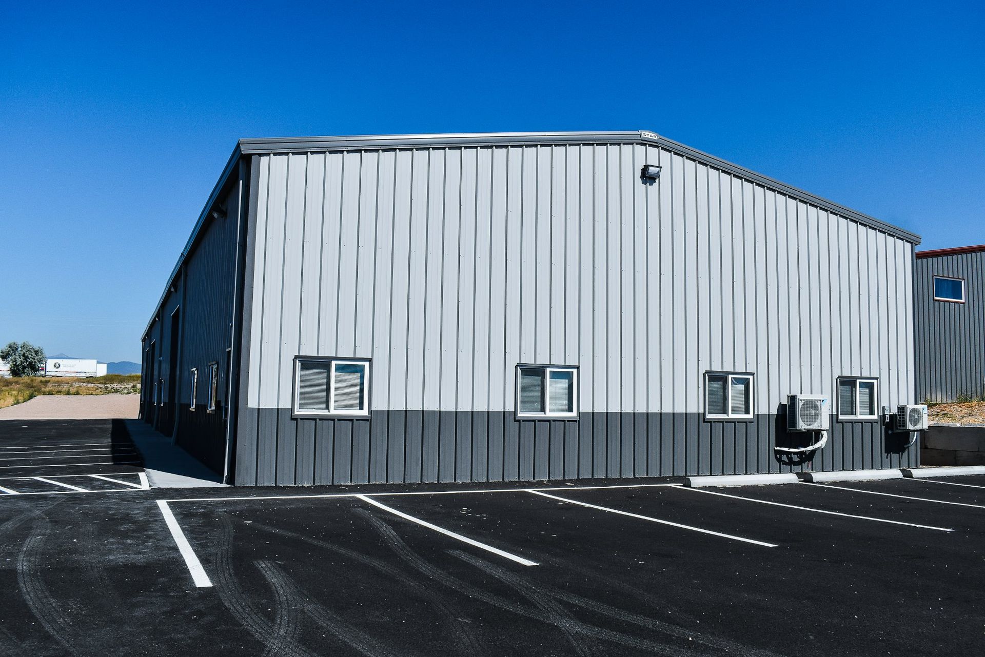 Gray metal warehouse building with small windows in a paved lot under a clear blue sky