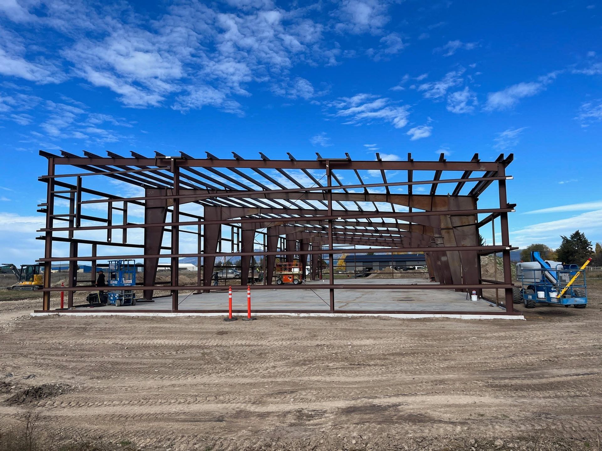 Steel-frame building under construction on a dirt lot under a bright blue sky.