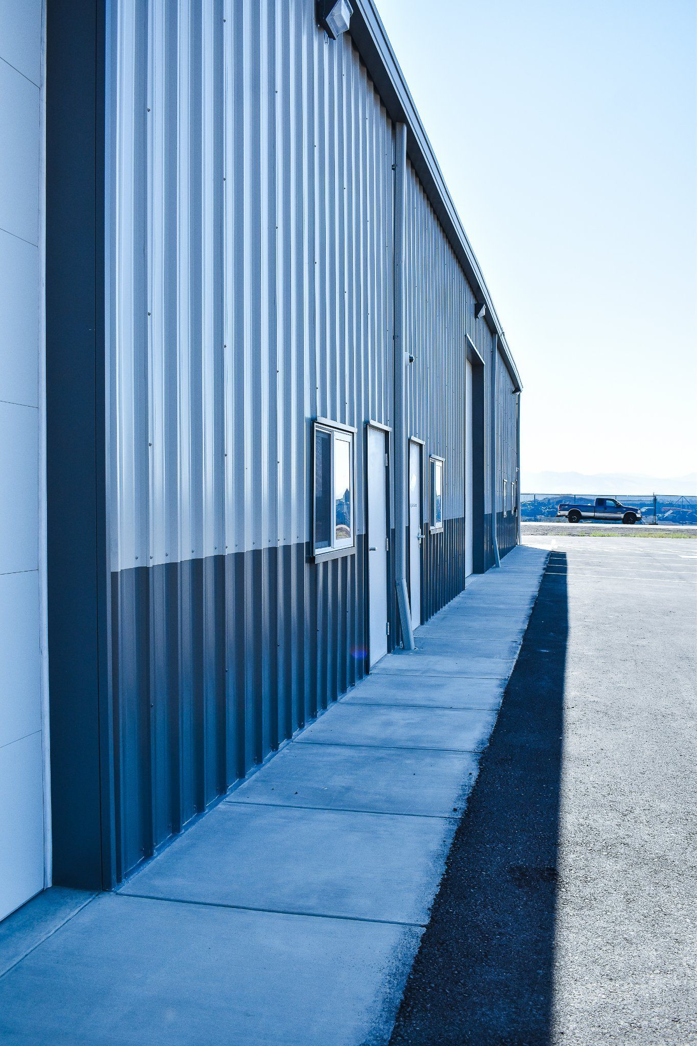 Blue metal warehouse wall with doors beside a concrete path and gravel lot under a bright sky