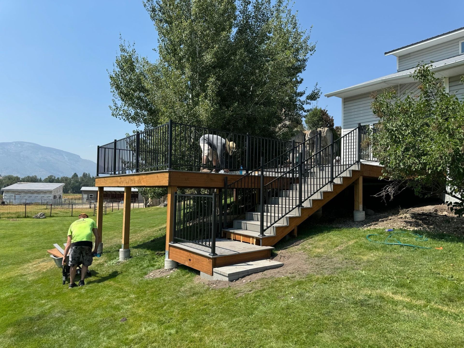 Person on a lawn mower near a raised wooden deck and stairs beside a house, with trees and mountains in the background