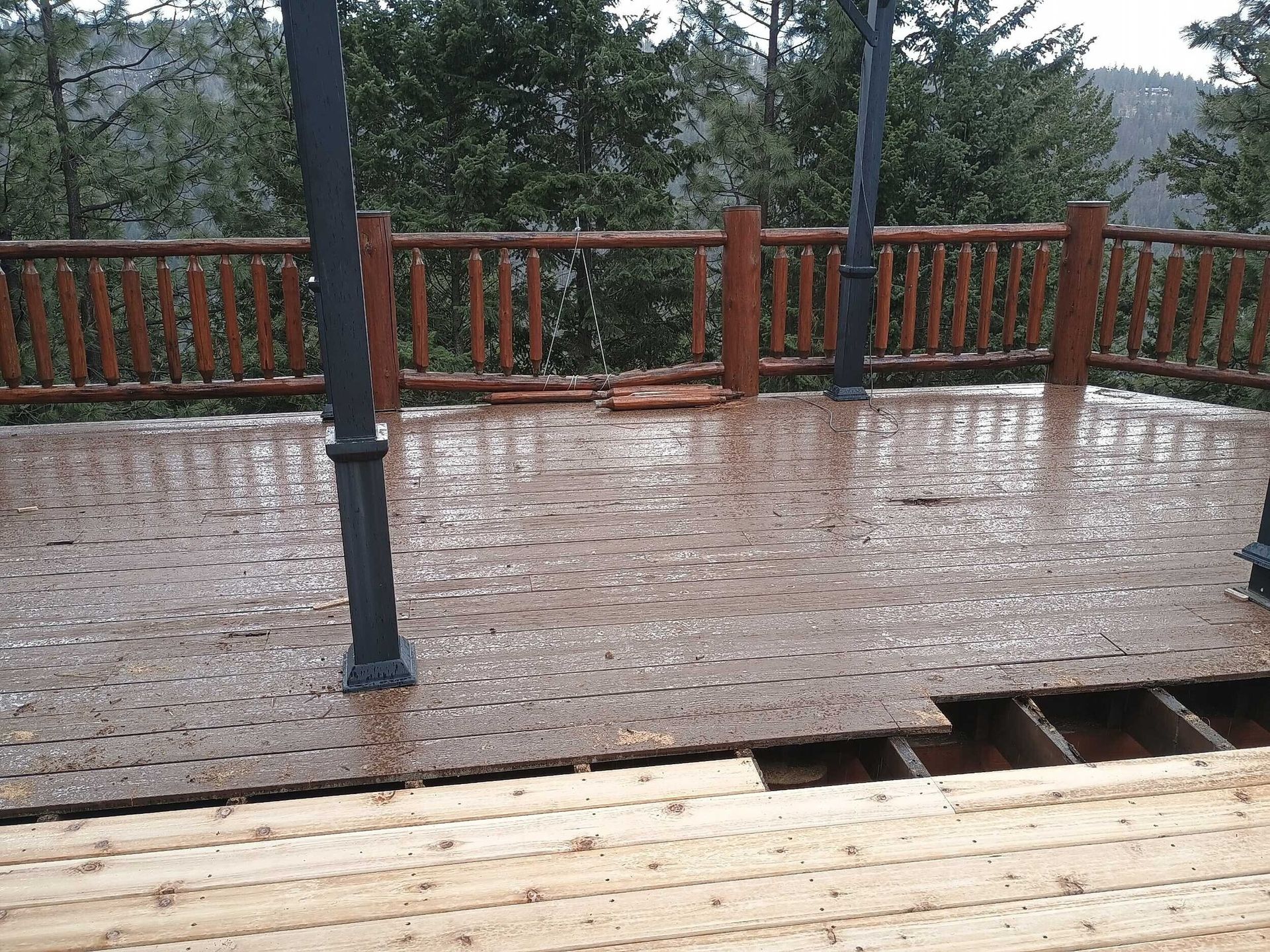 Rain-soaked wooden deck with railing and a missing section exposing beams, overlooking trees and hills
