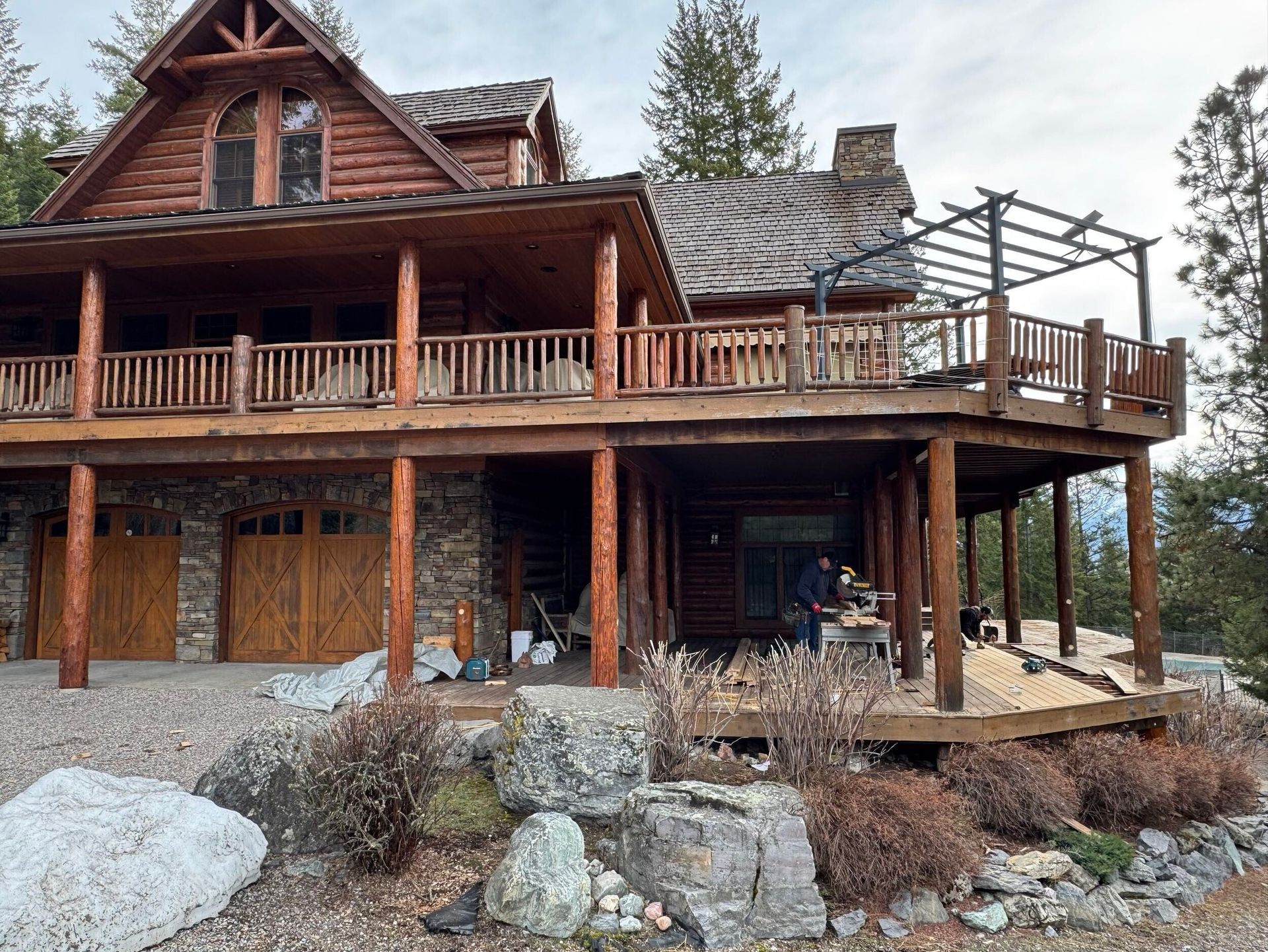 Rustic log cabin with wraparound porch, stone foundation, and gravel yard in a forest setting