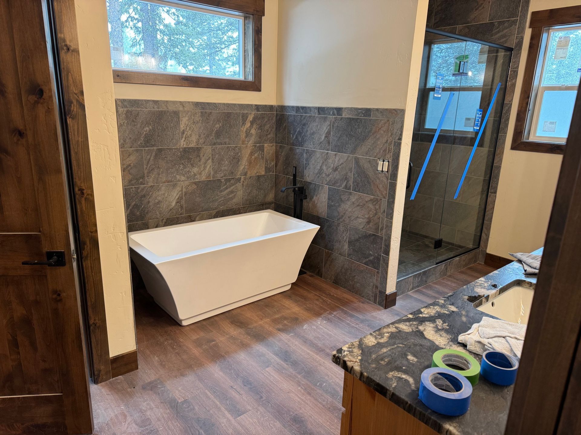 Bathroom with a freestanding white tub, dark tile walls, wood floor, and shower glass on the right.
