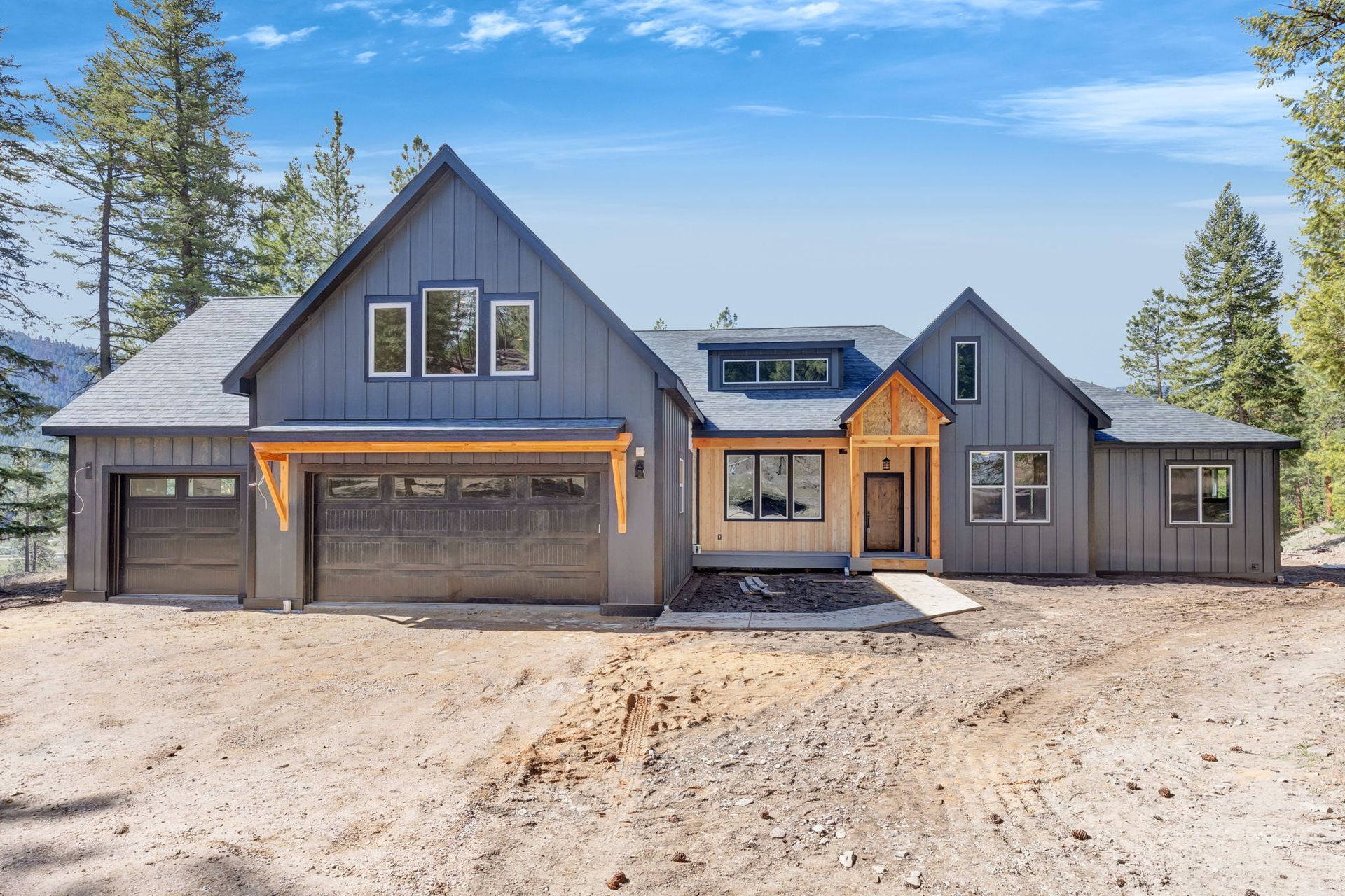 Modern gray house with wood accents, large garage, and a dirt front yard under a blue sky