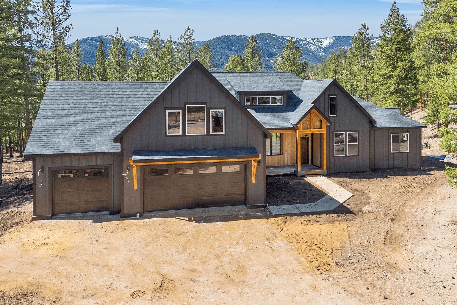 Dark gray mountain cabin with garage, surrounded by pine trees and mountains under a clear sky