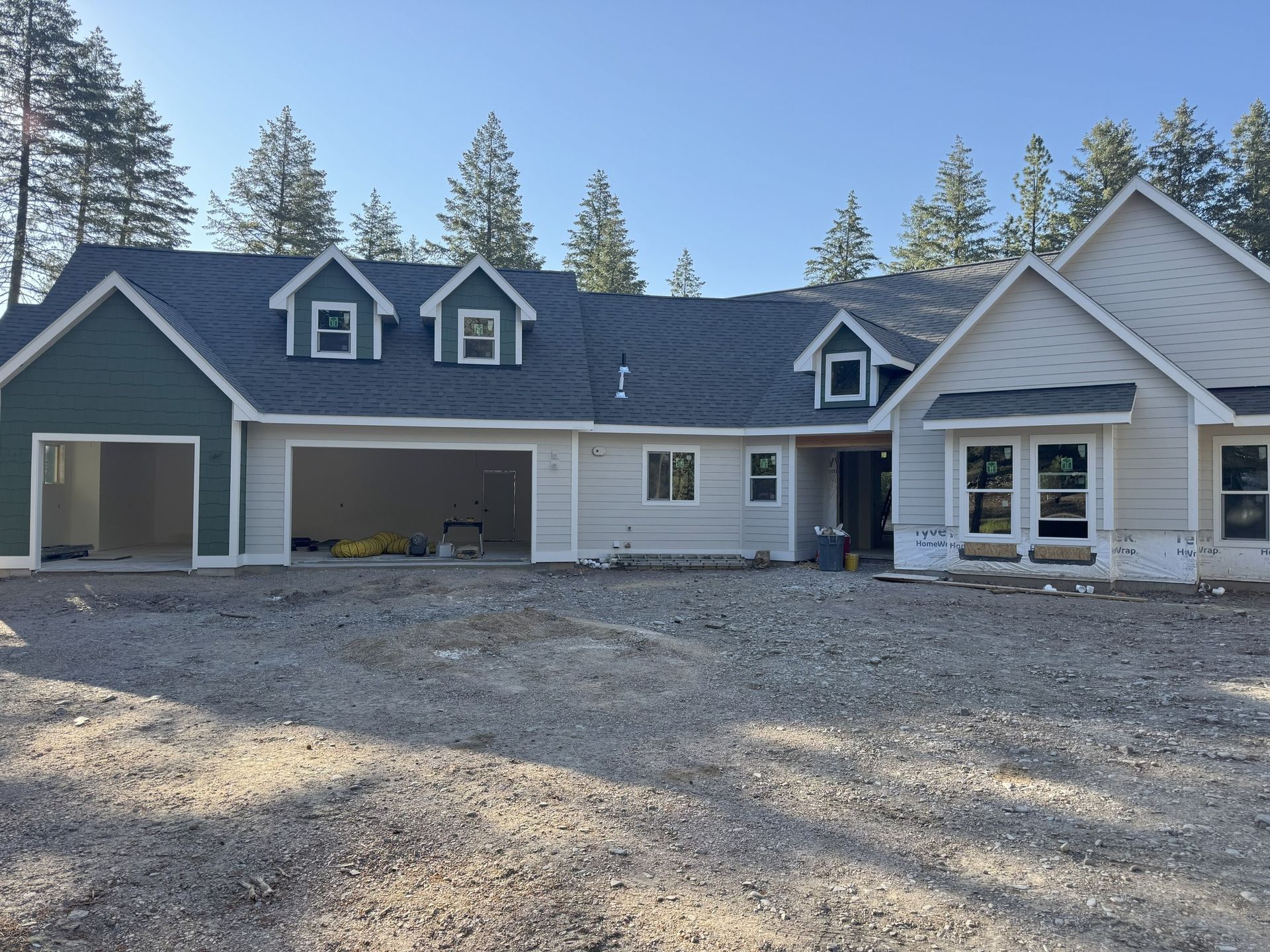 Large blue-gray house with white trim, attached garage, gravel driveway, and pine trees in the background