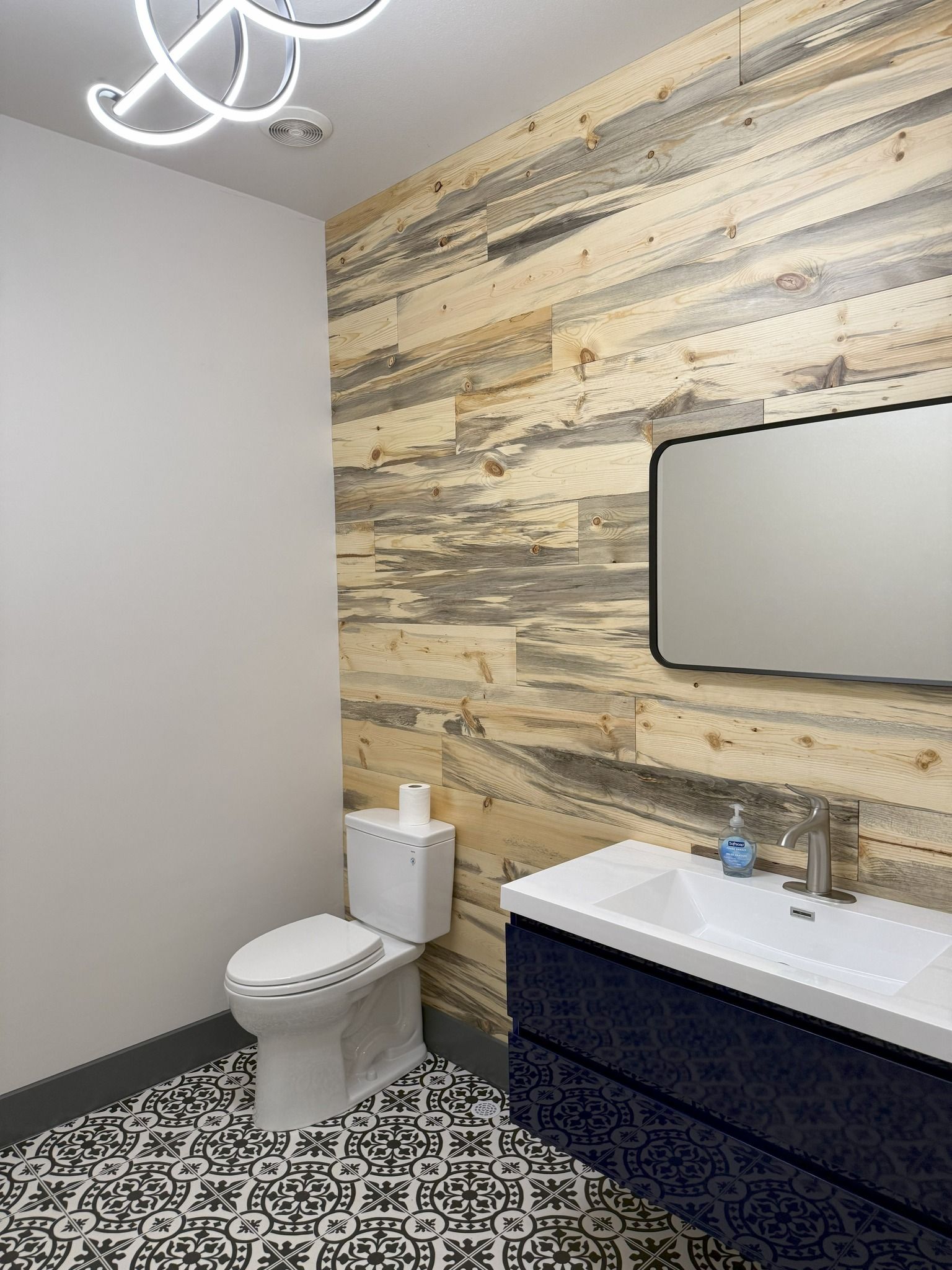Modern bathroom with patterned floor, wood accent wall, toilet, and vanity sink with mirror