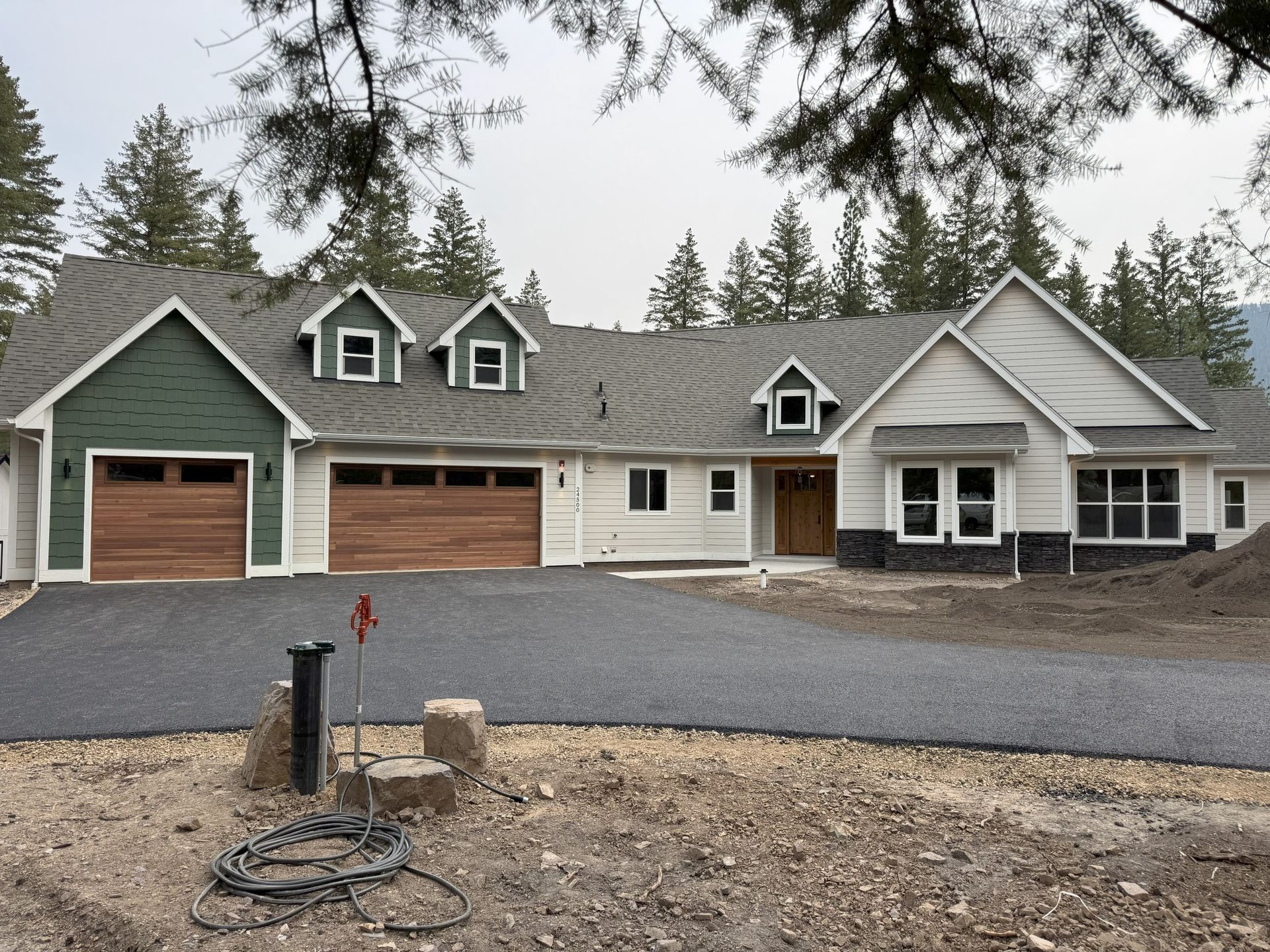 Large gray house with green trim, three wooden garage doors, and gravel driveway under overcast sky