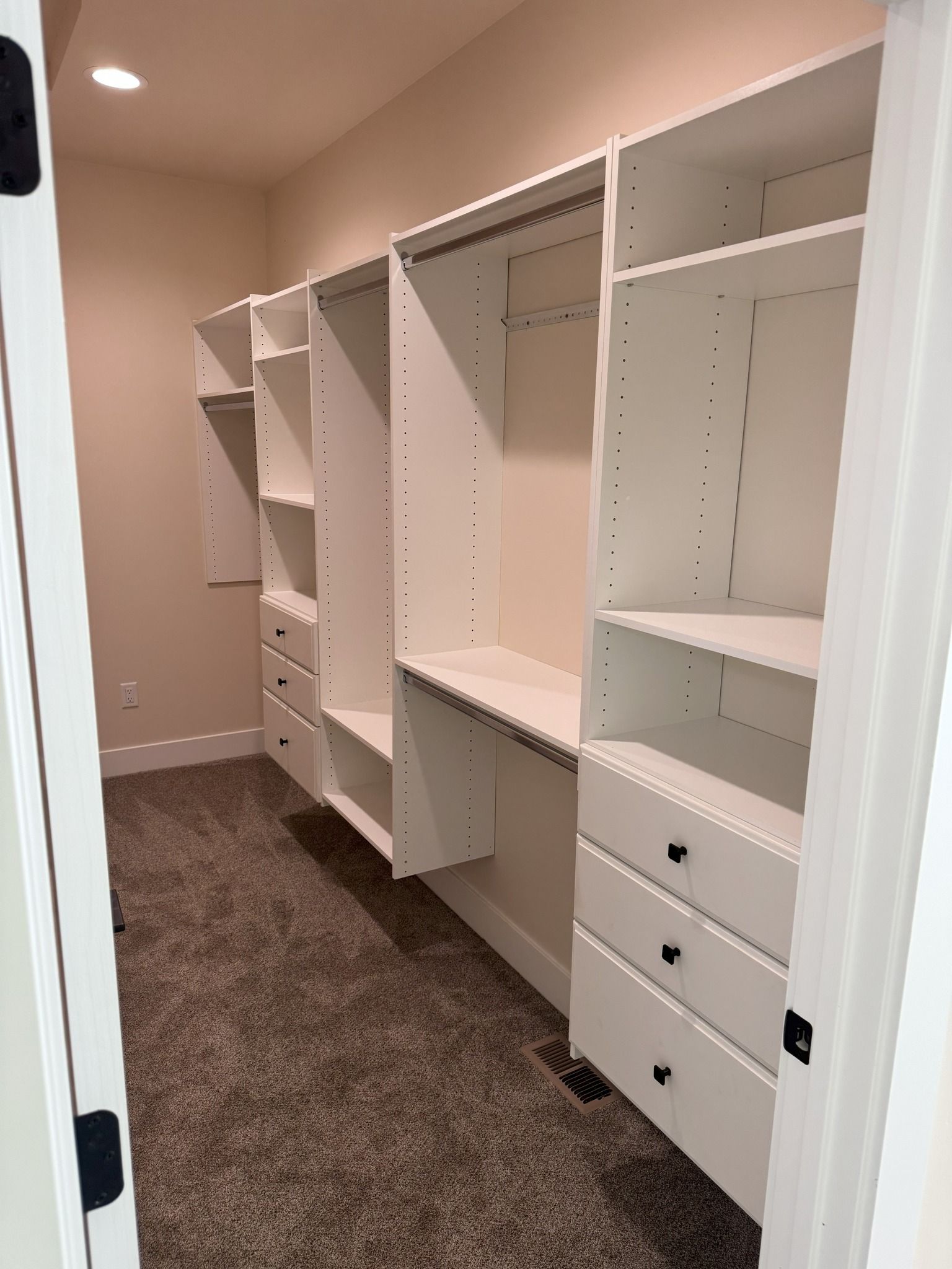 Walk-in closet with white shelving, drawers, and hanging rods on beige walls and carpet.