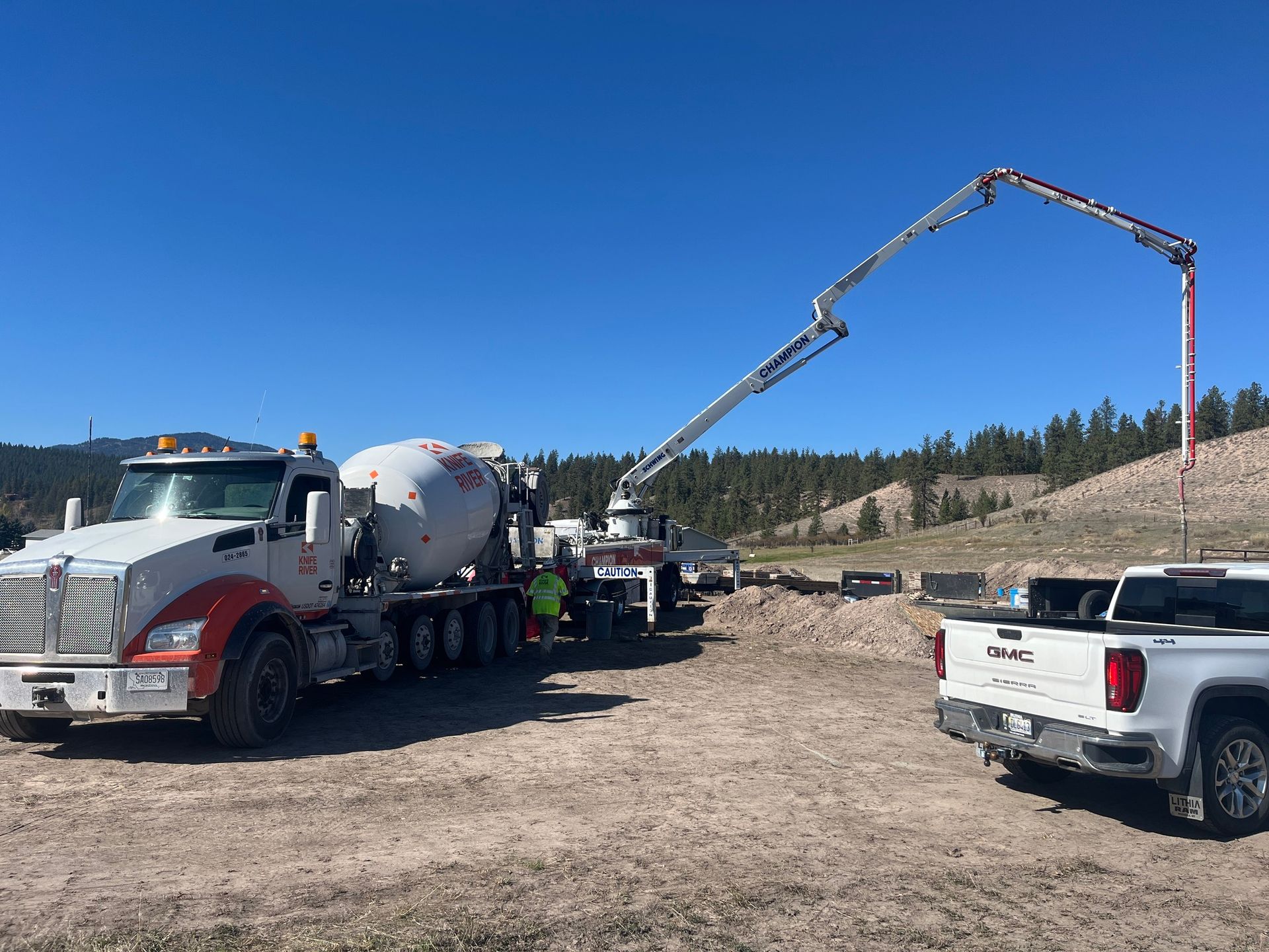 Concrete pump truck extending boom over a worksite with a pickup truck nearby under a clear blue sky