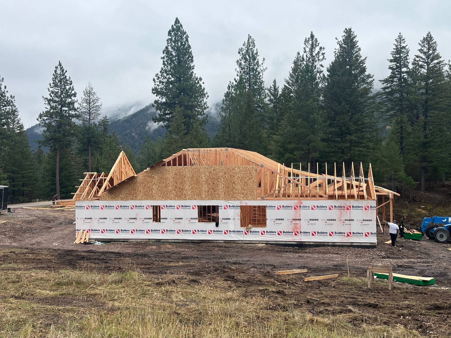 House under construction with framed roof on a rural lot, surrounded by tall pine trees and mountains