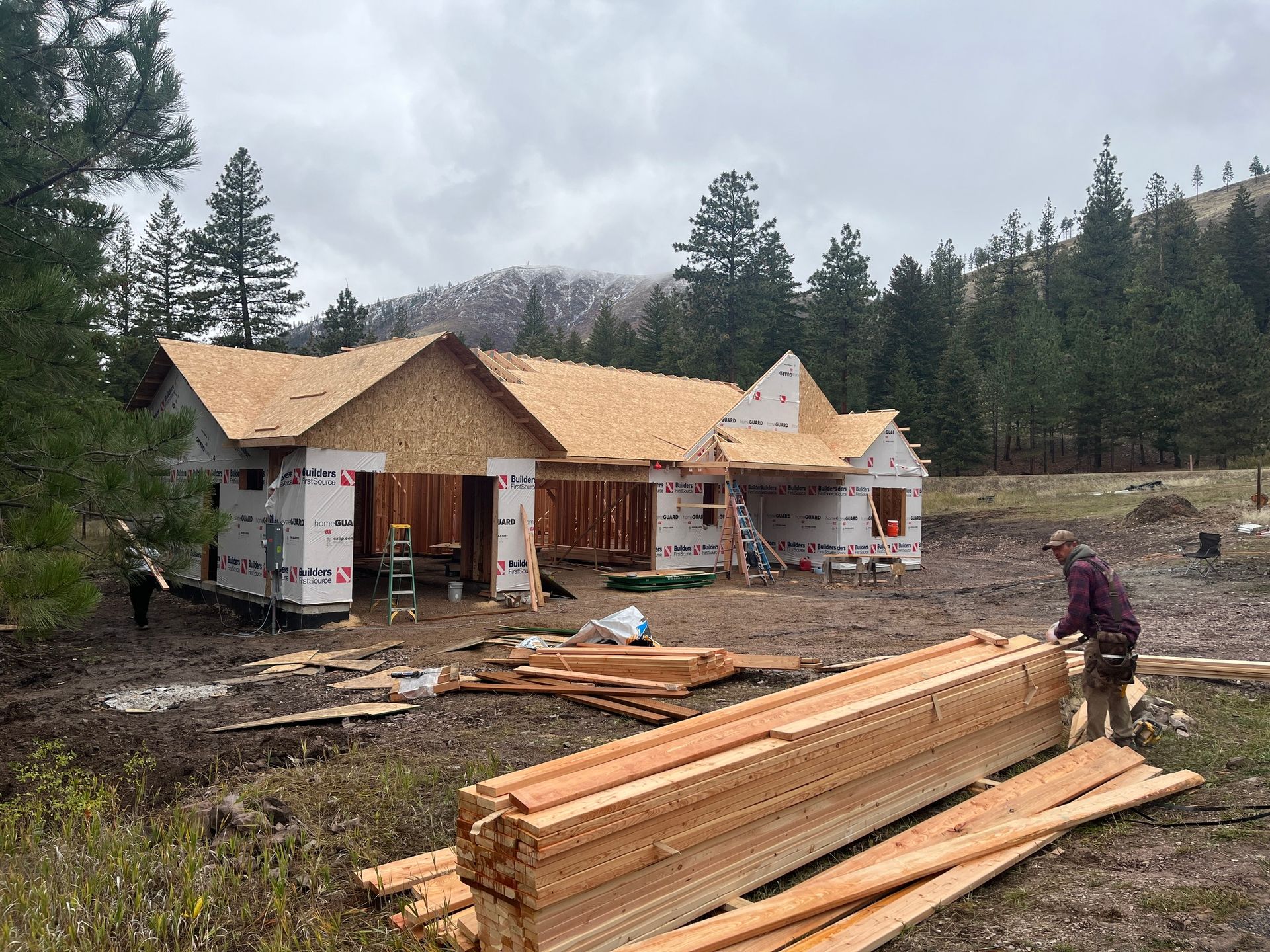 Framed wooden house under construction in a muddy forest clearing, with lumber stacks and workers nearby