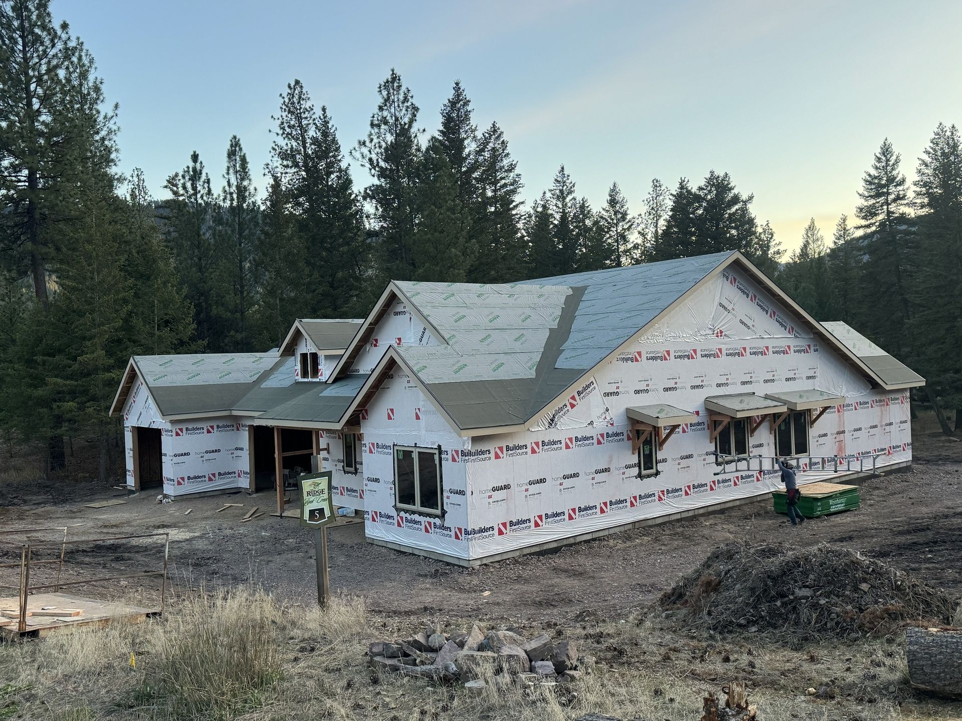 Partially built house wrapped in housewrap, surrounded by trees and construction dirt at dusk