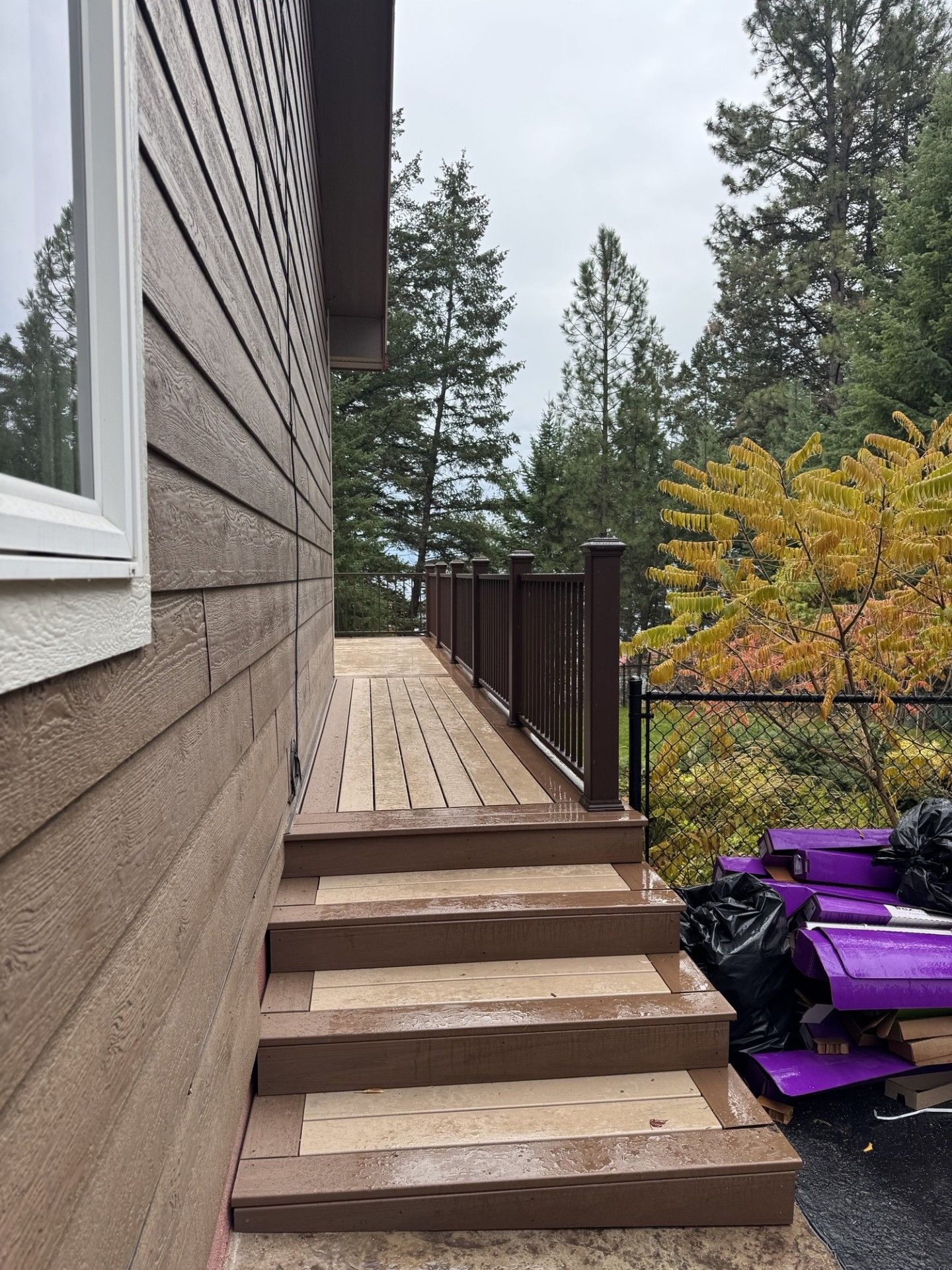 Side view of a house deck with wooden steps, dark railing, and trees in the background.