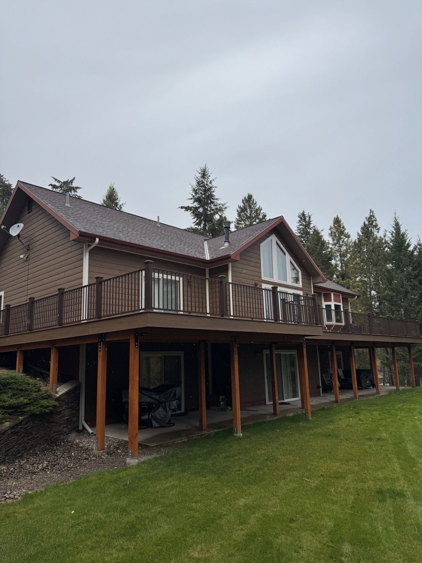 Wooden house with a wraparound porch and upper balcony beside a grassy yard and trees